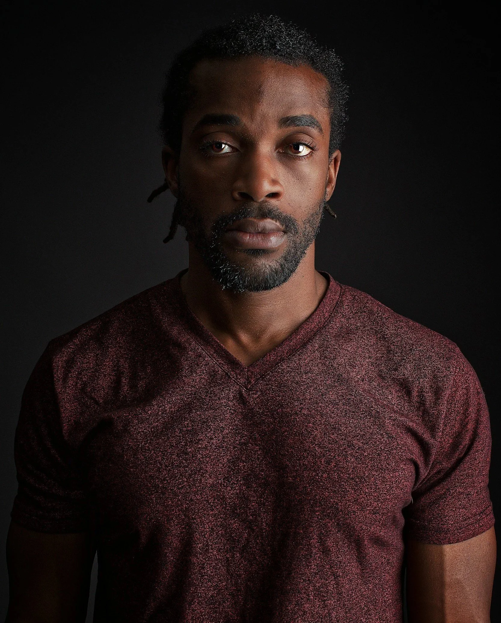 Portrait of a Black man with dreadlocks, wearing a maroon V-neck shirt, against a black background.