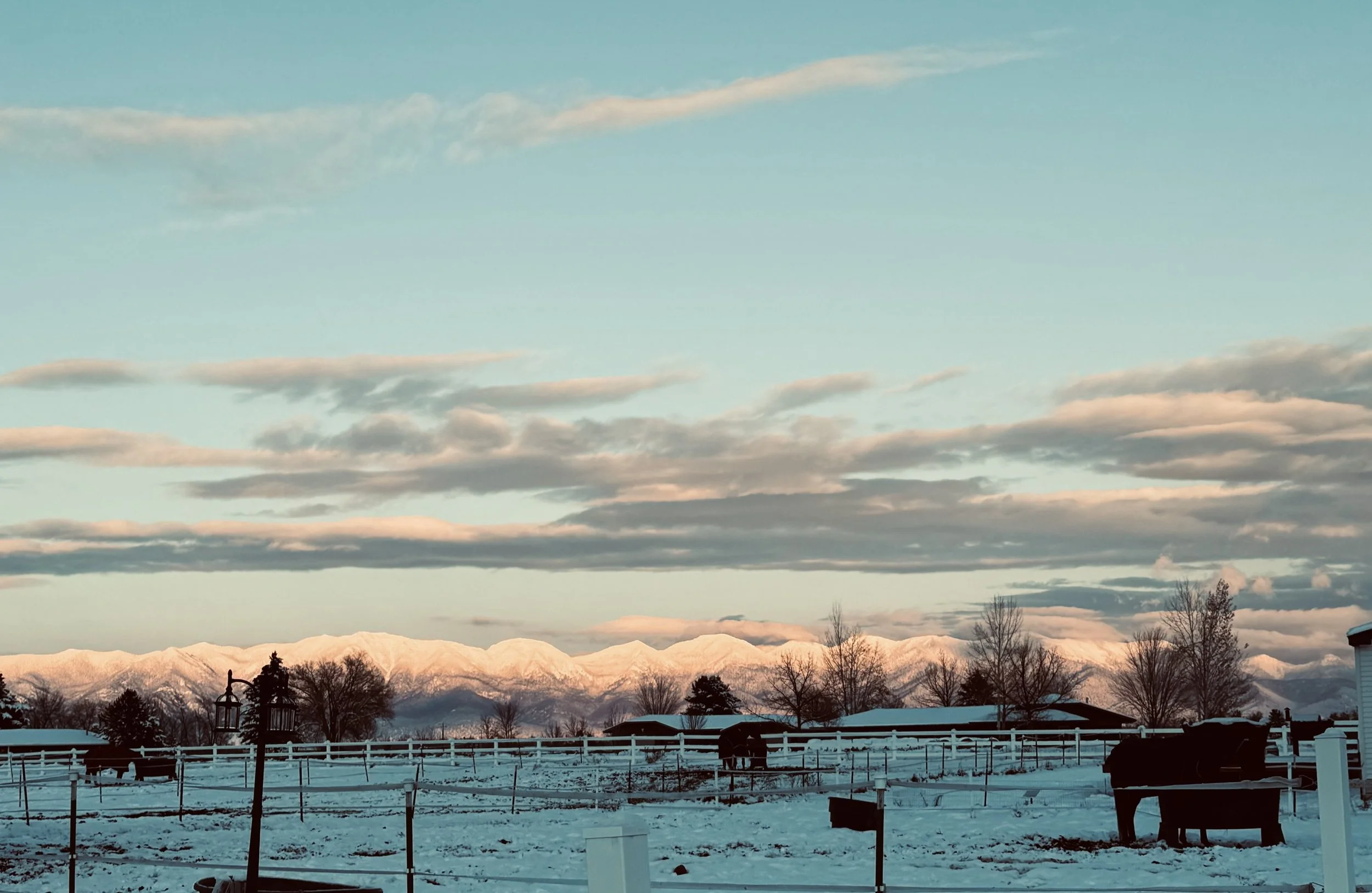 Snow-covered landscape with fenced fields, trees, and distant snow-capped mountains under a sky with scattered clouds.