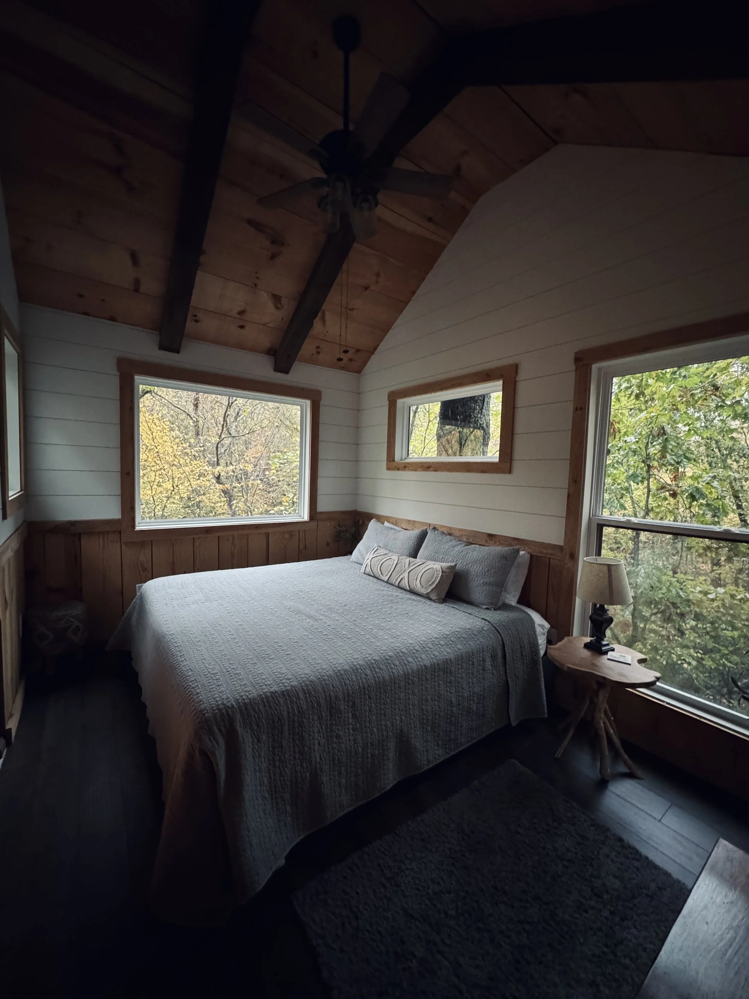 Cozy bedroom with a bed featuring a white textured blanket, two gray pillows, and a decorative pillow, wooden wall paneling, large windows showing trees outside, a small wooden side table with a lamp, and a dark carpeted floor.