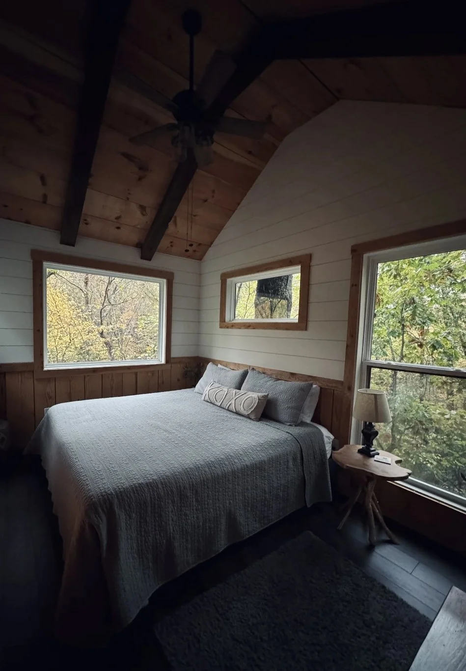 A cozy bedroom with a large bed covered in a quilt, three pillows, and a small decorative cushion. There are two large windows showing trees outside, and a wooden side table with a lamp on it next to the bed. The ceiling is vaulted with wooden beams and a ceiling fan.