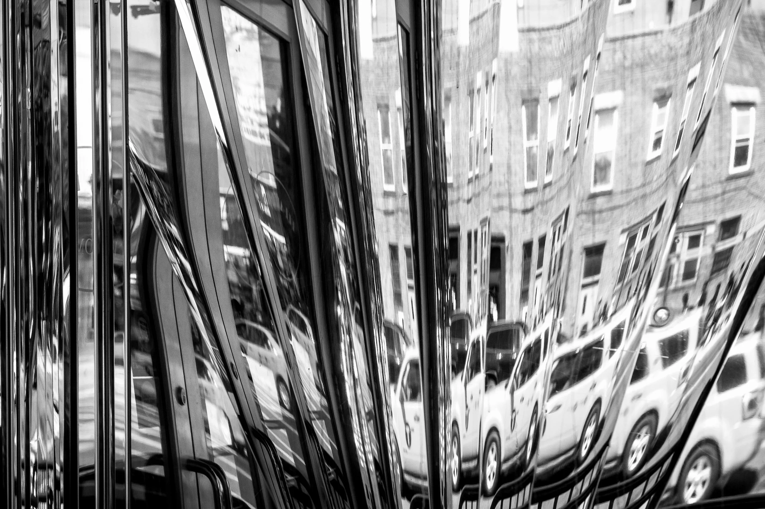 Black and white photo of a highly reflective, curved metal surface distorting reflections of parked cars and buildings.