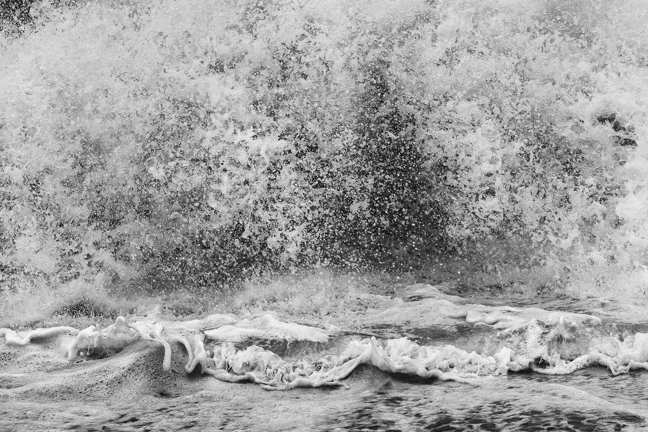 Black and white photo of ocean waves crashing against the shore, with spray and foam in the air.
