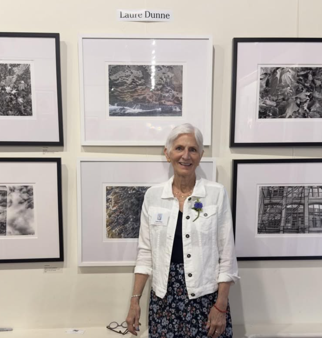 An elderly woman standing in front of framed black and white photographs at an art exhibition, smiling at the camera.