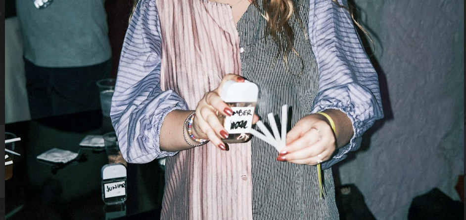 Person holding a small bottle with a label that says 'NUMBER' in a party scene, with other people and drinks visible in the background.