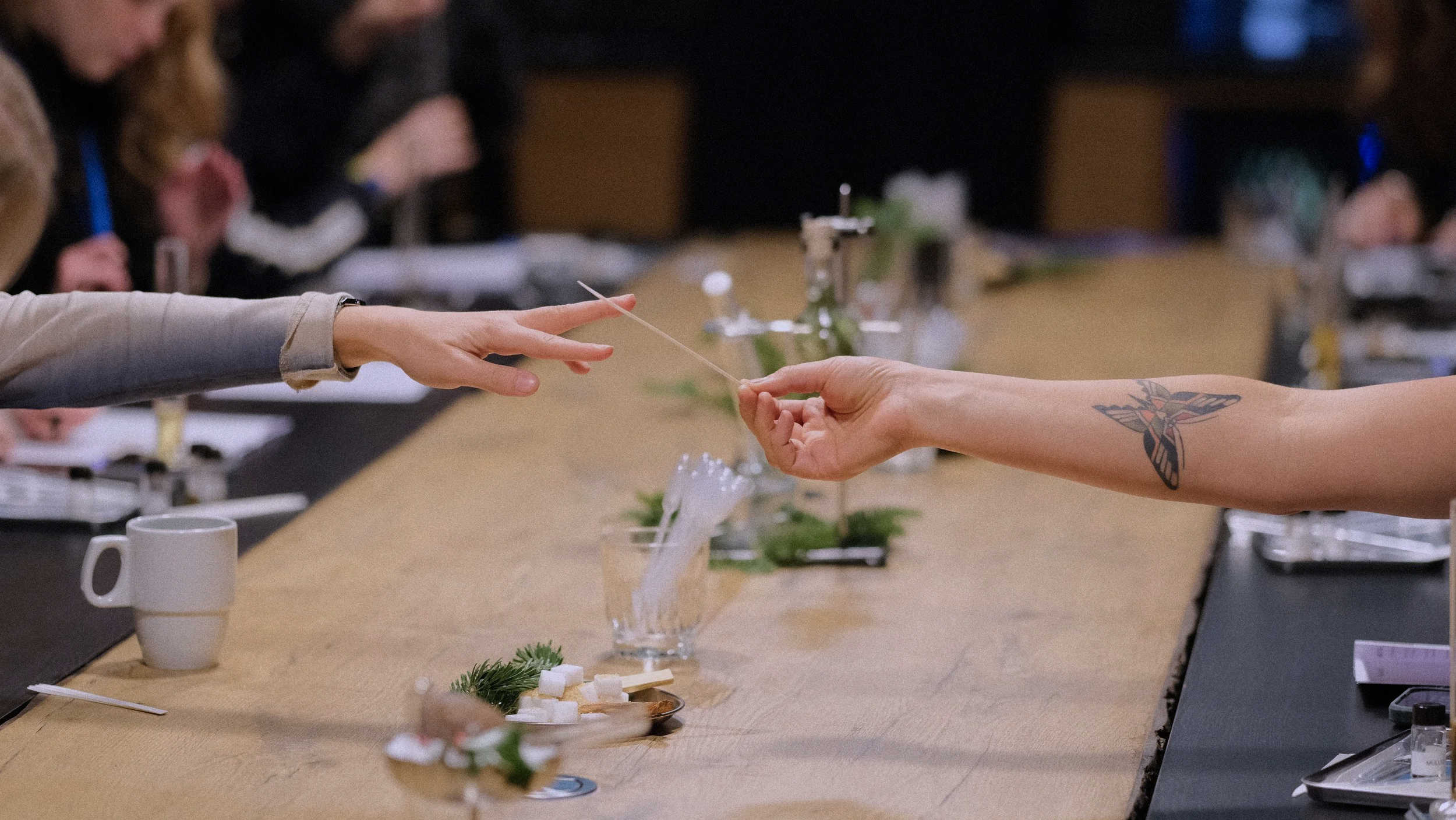 A person with a tattoo on their arm receives a fragrance test strip from another person during an olfactory workshop, with various supplies on the table.