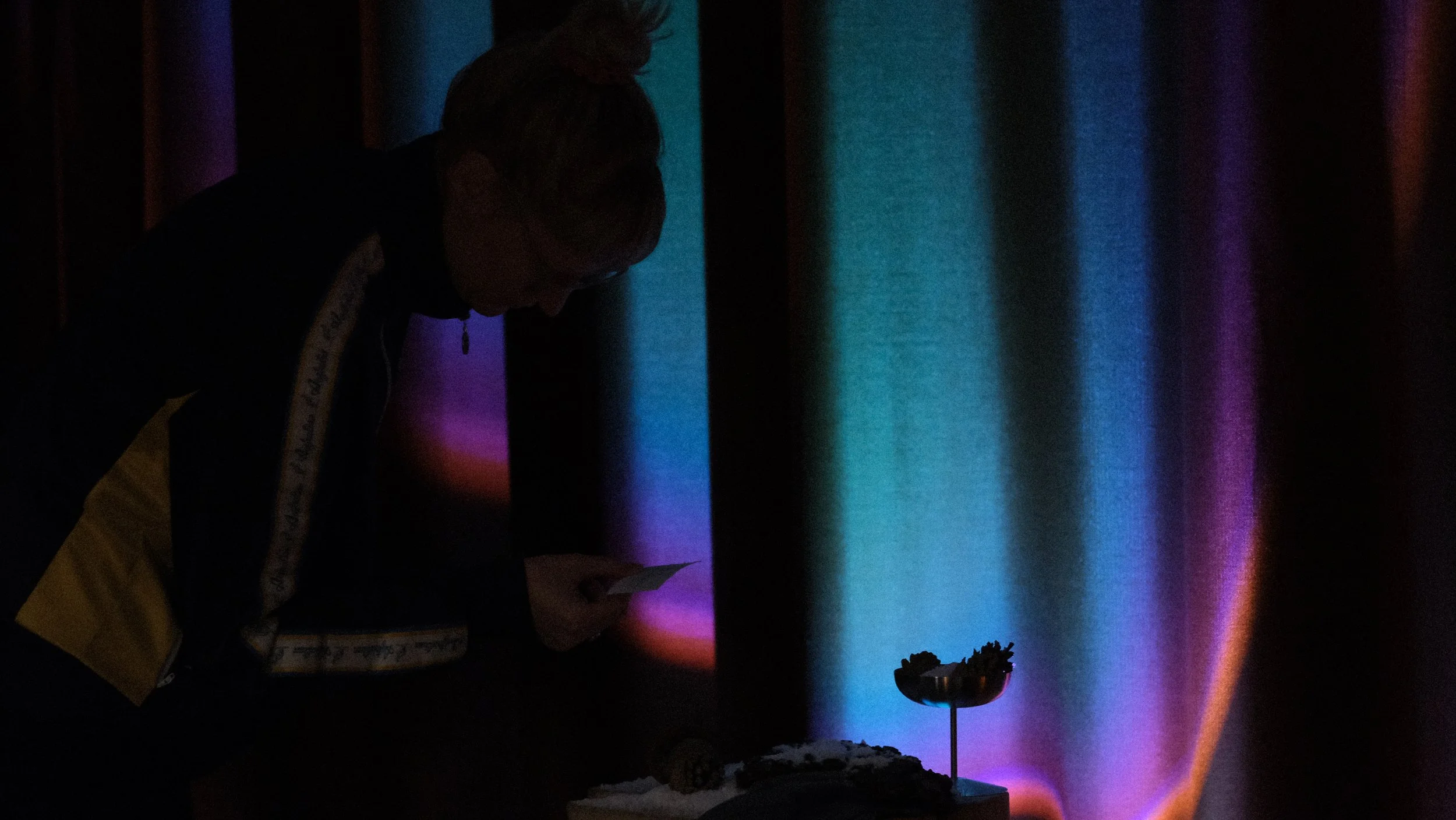 A person on stage looking at a small object while holding a piece of paper, with a colorful curtain in the background and a small sculpture on a stand in front of them.