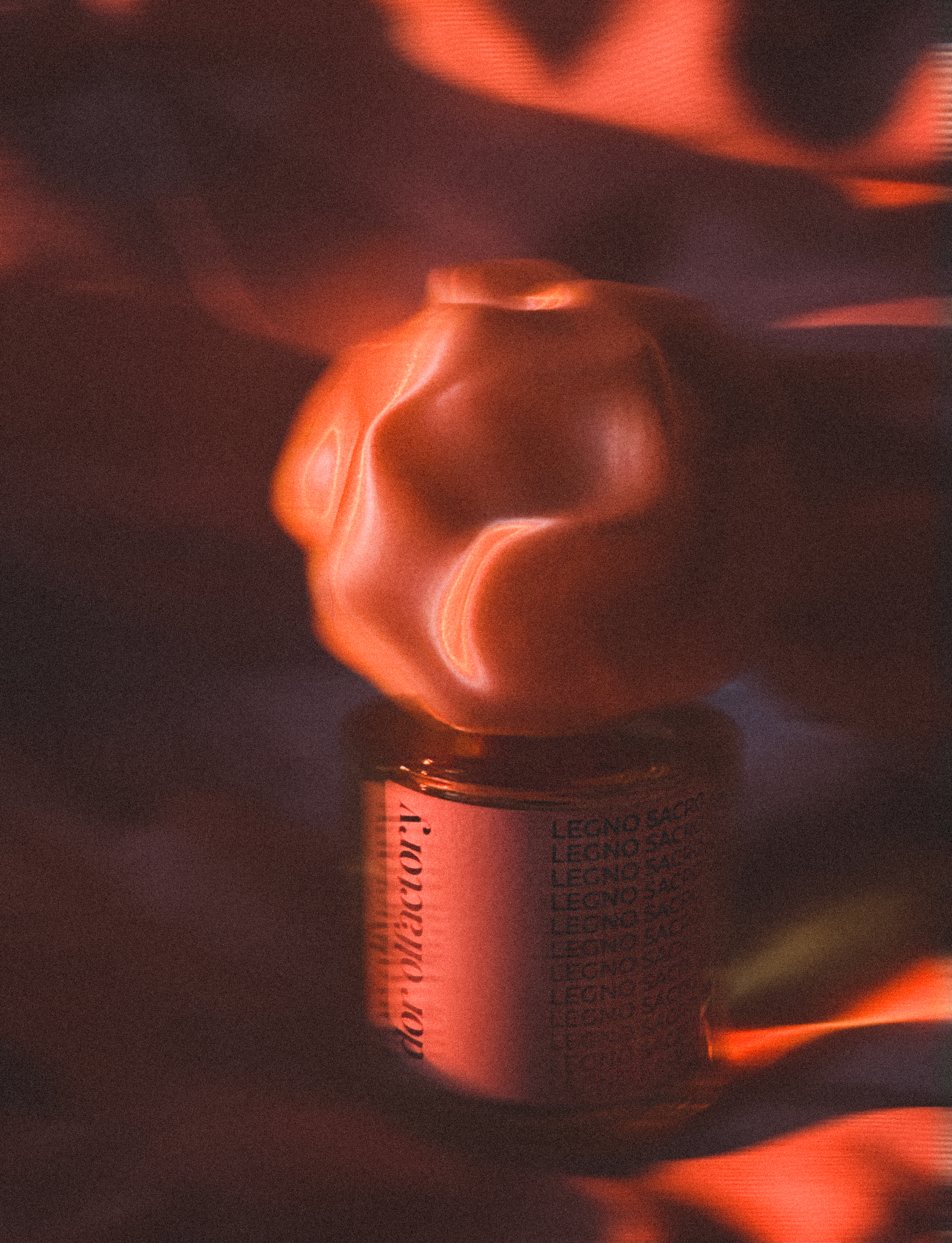 A small perfume bottle 'LEGNO SACRO' with an orange-red light reflecting on its surface, set against a dark background with similar glowing reflections.