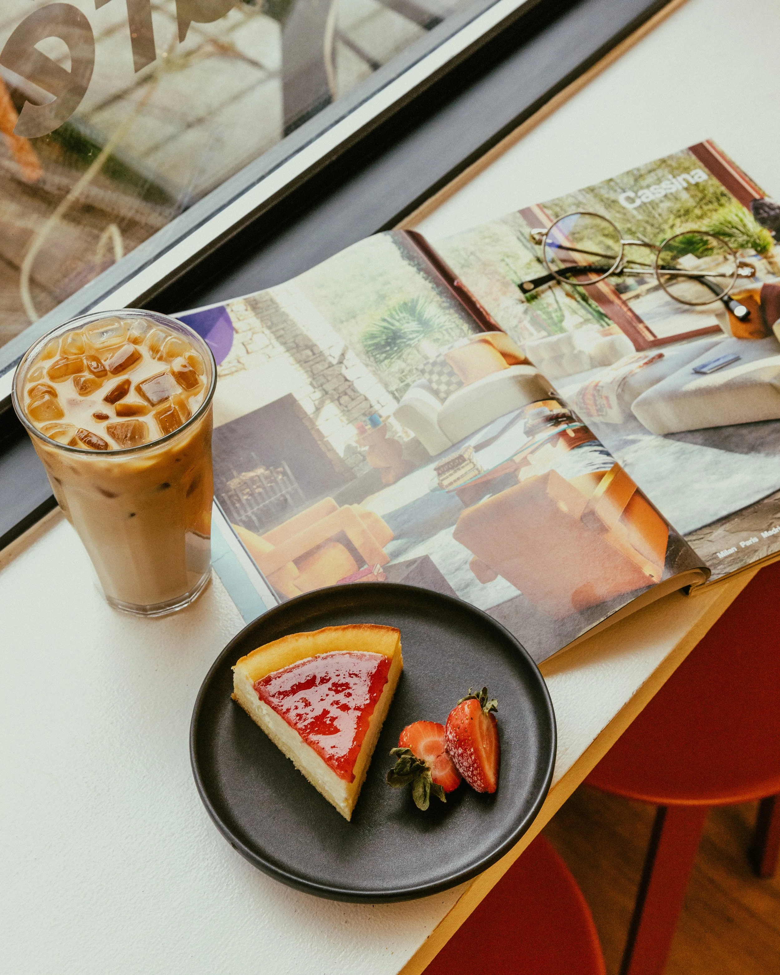 A table with a glass of iced coffee, a slice of strawberry cheesecake, and two strawberries on a black plate. An open magazine and a pair of round glasses rest on the table, with sunlight coming through a nearby window.