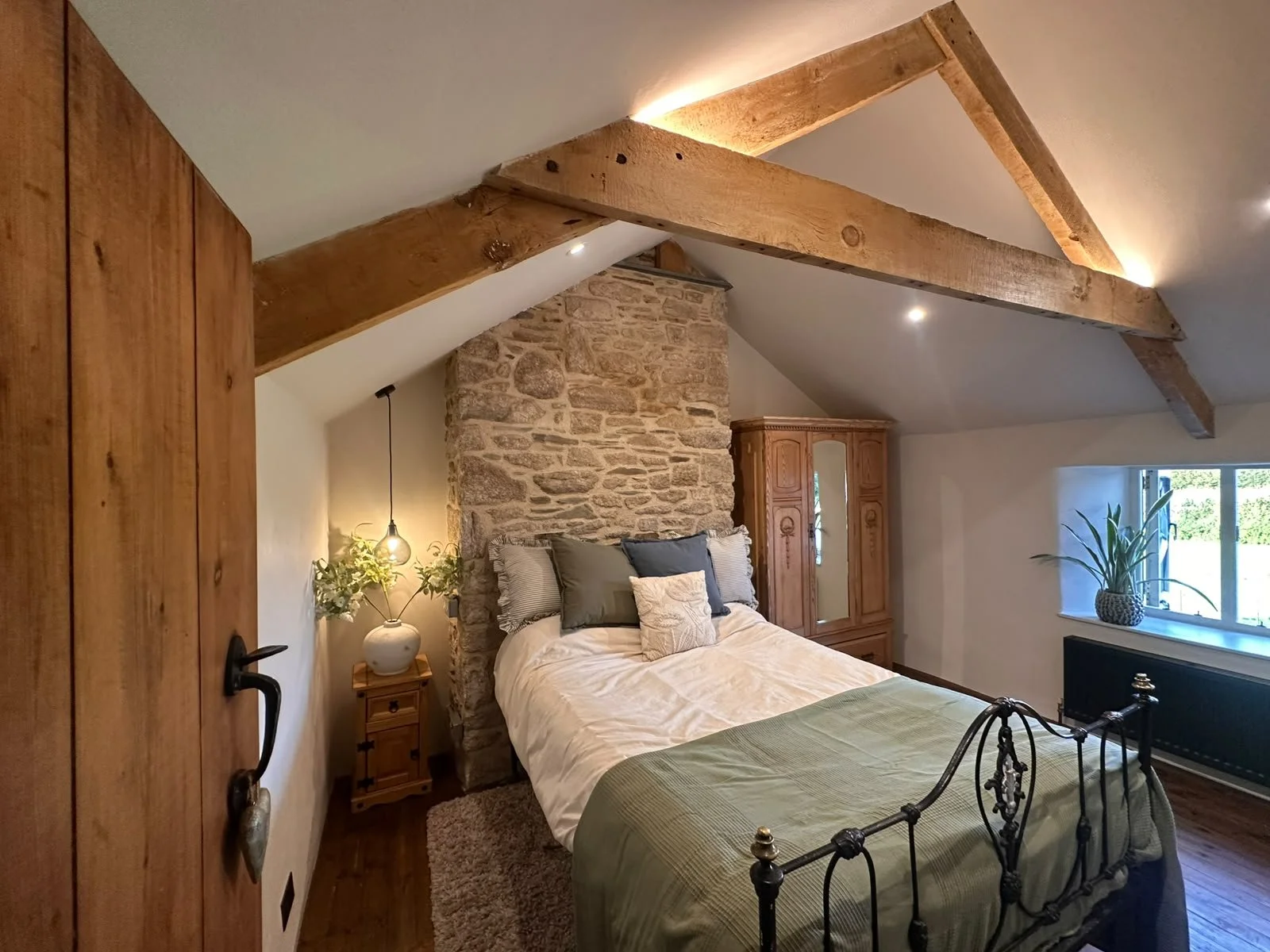 A cozy bedroom with a sloped ceiling featuring exposed wooden beams, a stone accent wall behind the bed, a black metal bed frame with beige and gray pillows, a green bedspread, a wooden wardrobe, a small wooden nightstand with a potted plant and a ha