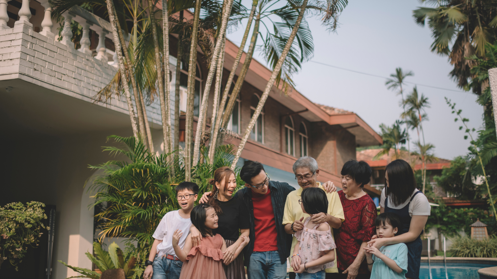 Family and friends laughing and talking outdoors in a backyard with lush greenery and a swimming pool.