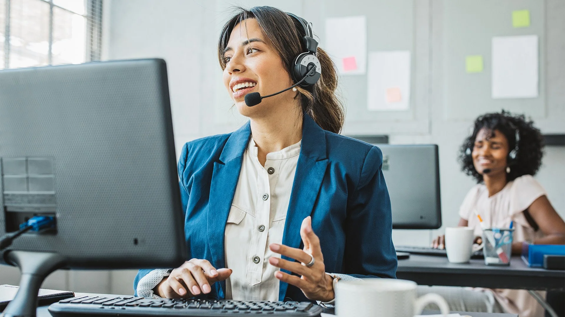 Two women wearing headsets, working in an office with computers and notebooks, smiling and engaging in a call.