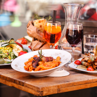 Food and drinks on a wooden table, including a bowl of spicy shrimp, a plate of salad, a meat dish, and glasses of red and orange beverages.
