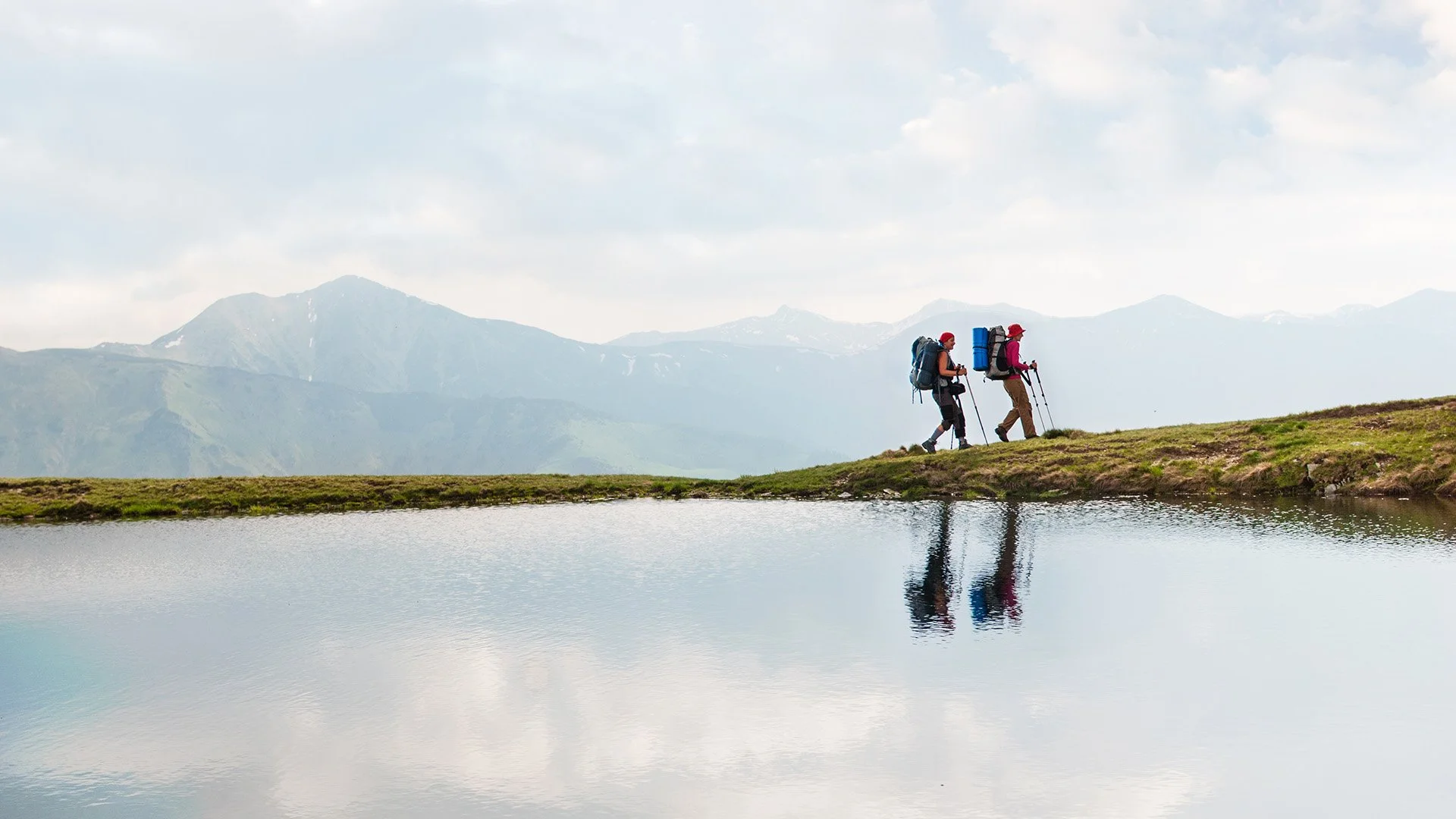 Two hikers with backpacks and trekking poles walking along a grassy trail next to a reflective body of water in a mountainous landscape.