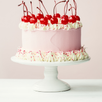 A pink and white layered cake decorated with whipped cream, colorful sprinkles, and topped with cherries.