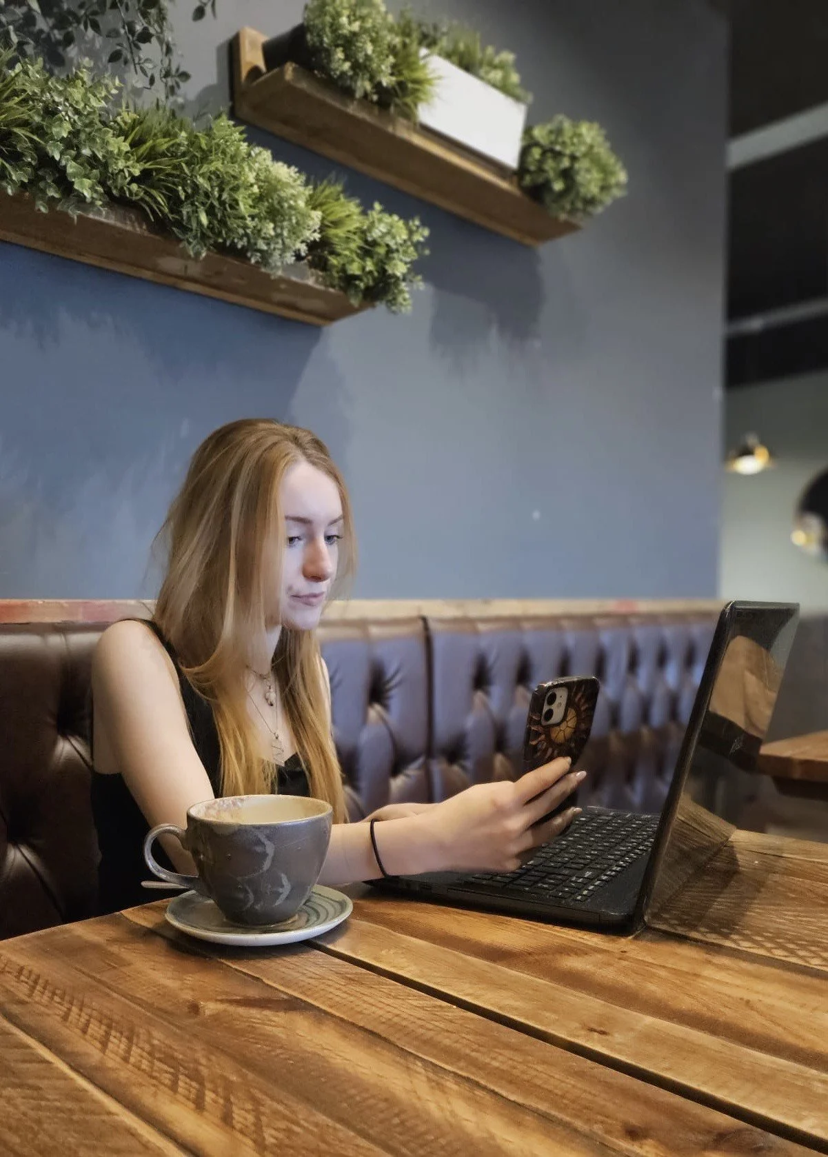 A young woman with long red hair sitting at a wooden table in a coffee shop, looking at her phone and working at her laptop. There is cup of coffee on a saucer in front of her, and plants in the background.