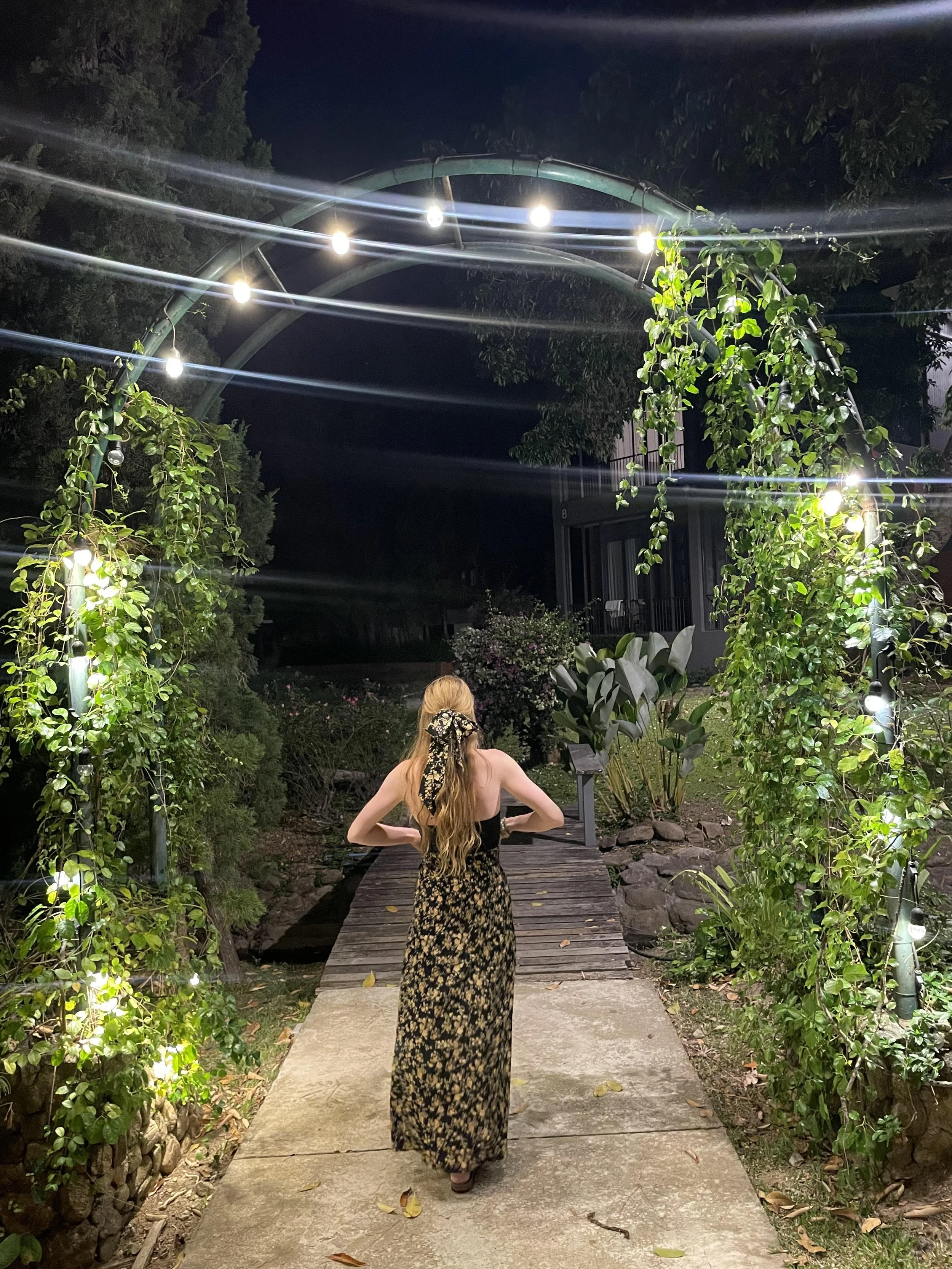 Woman with long hair wearing a black dress with a floral pattern, standing on a garden path at night under an archway decorated with string lights and green vines.