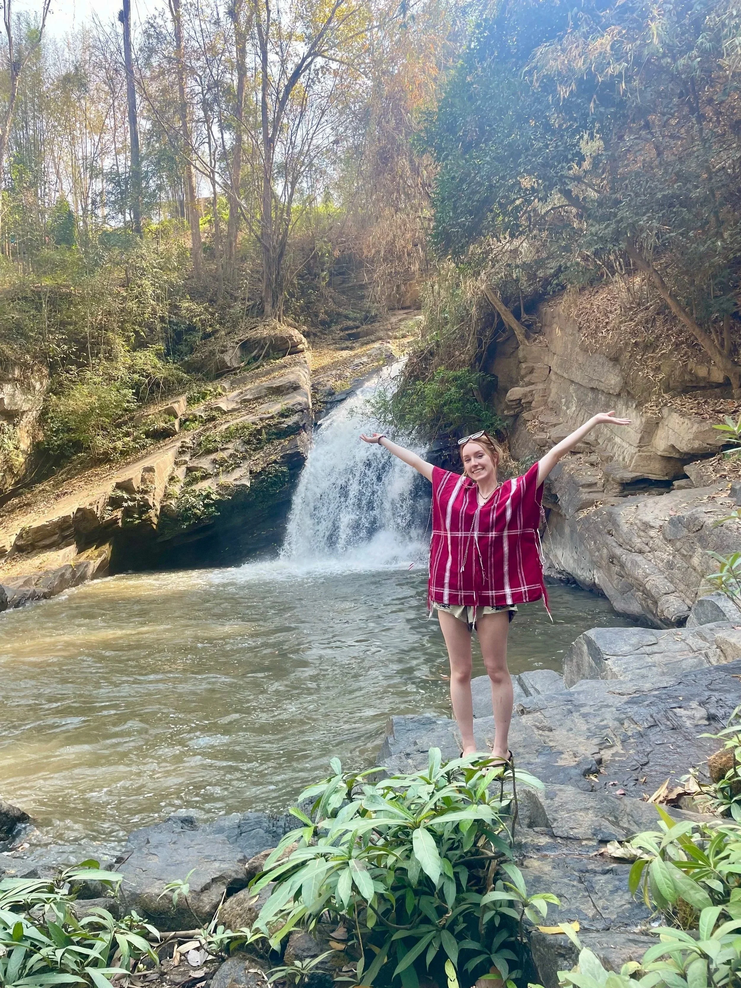 Zoe stands on rocks with arms outstretched near a small waterfall and stream in a forested area in Chiang Mai Thailand.