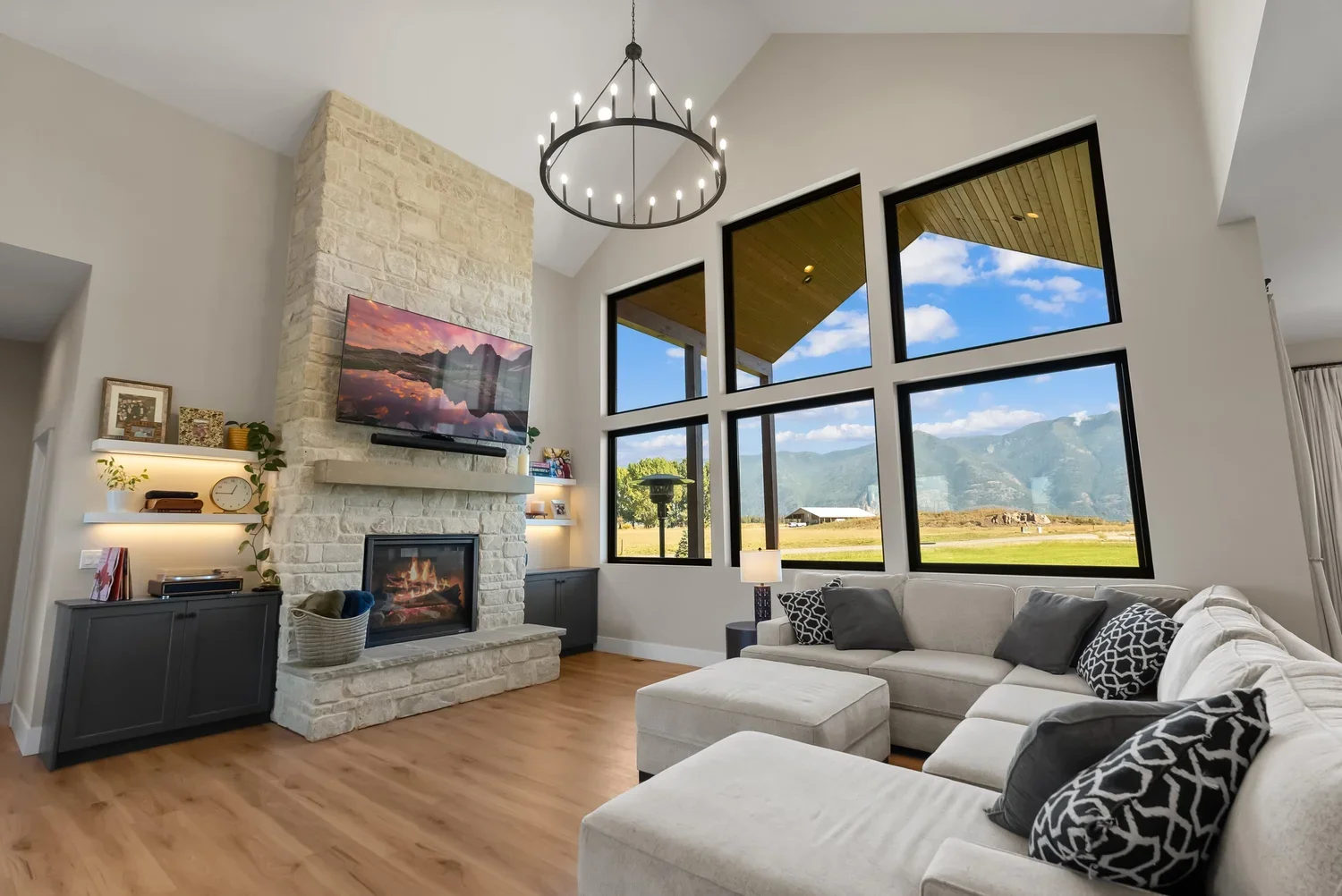 Modern living room with large windows showcasing mountain views, a high ceiling with a chandelier, a stone fireplace with a mounted TV, and a white sectional sofa with black and white patterned pillows.