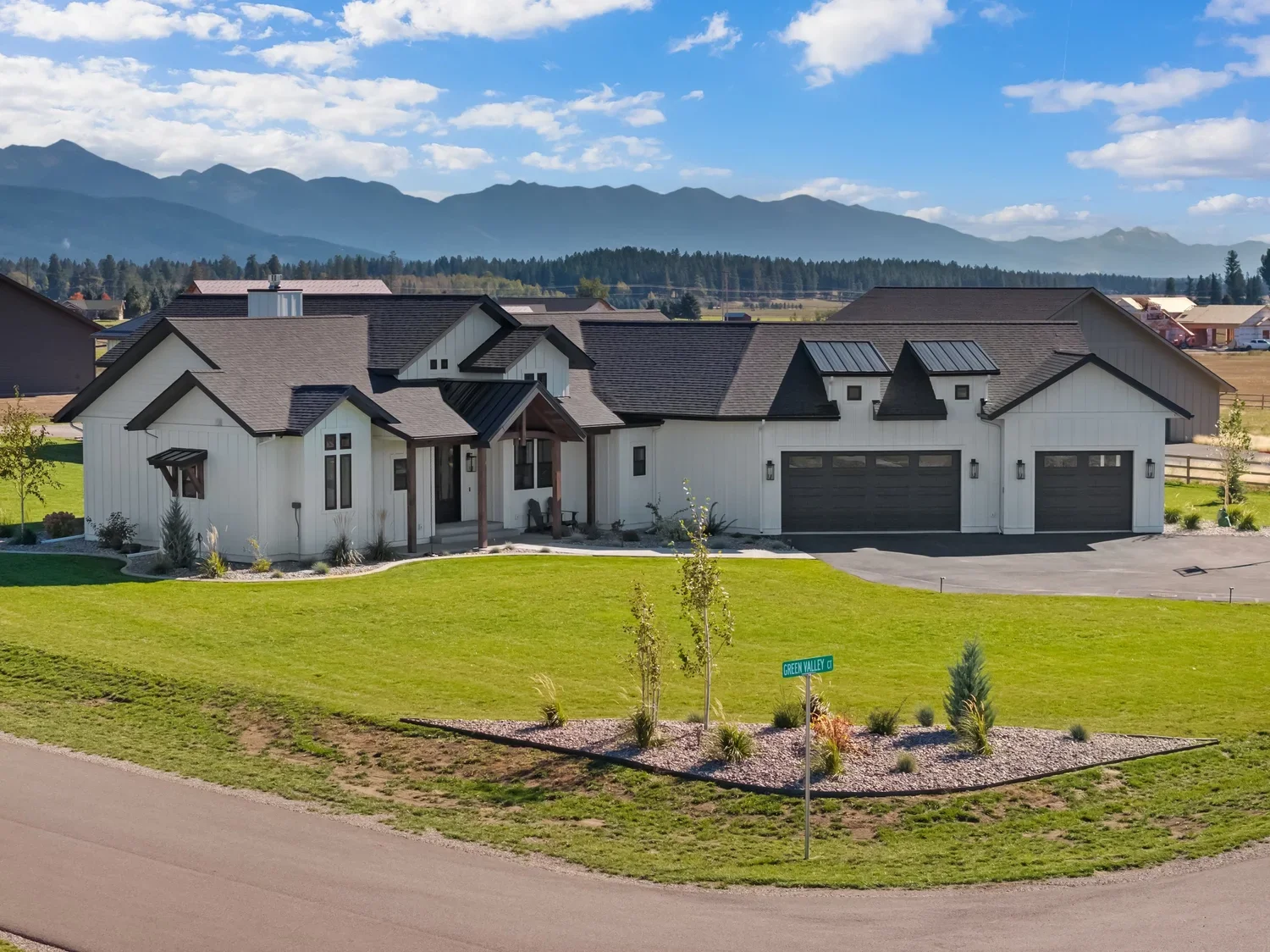 Modern single-story house with a white exterior, dark roof, and attached garage, surrounded by a landscaped front yard with small trees and bushes, located in a suburban neighborhood with mountains in the background.