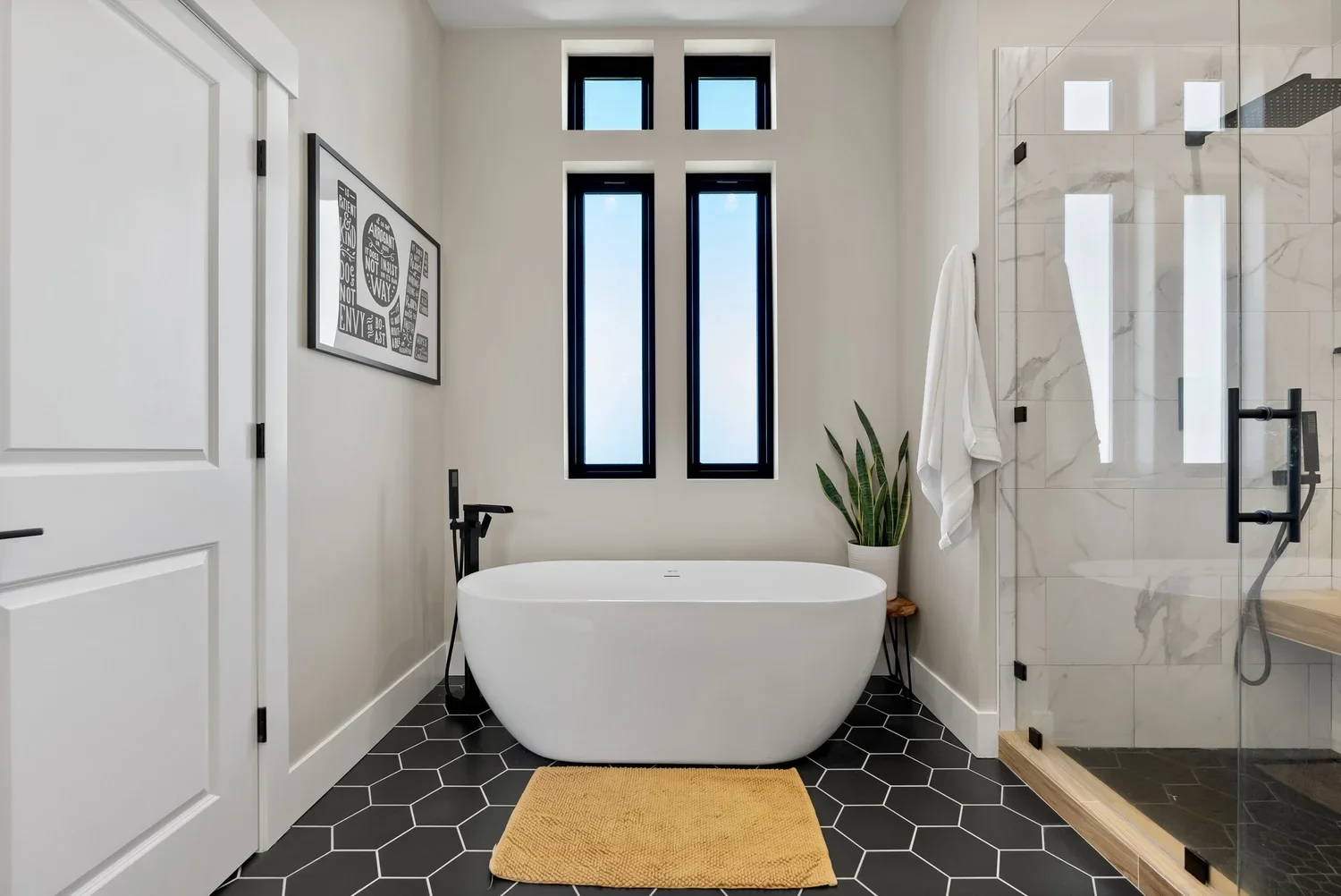 Modern bathroom with black hexagon tile flooring, a white freestanding bathtub, tall narrow windows, and a glass shower enclosure. Features include a plant, towel, art on the wall, and a yellow bath mat.