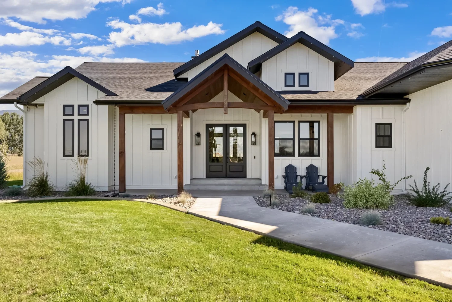 Front view of a mountain modern house with white board and batten siding, dark trim windows and a dark front door, wooden porch with two chairs, well-maintained lawn, and a clear blue sky.
