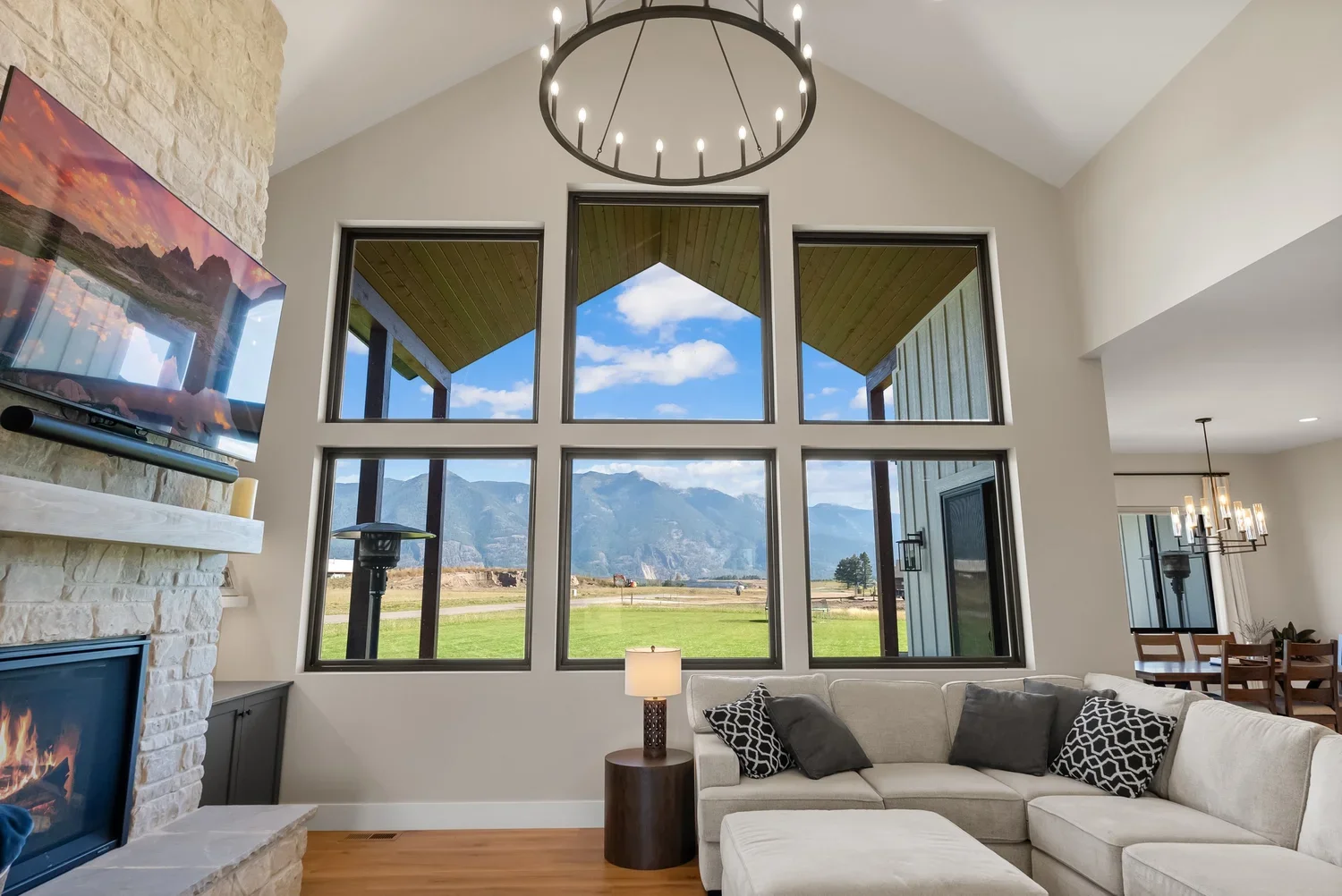 Interior of a living room with large windows showing mountains and blue sky outside, a white sectional sofa with black and white pillows, a side table with a lamp, television above a stone fireplace, and a dining area in the background with a chandel