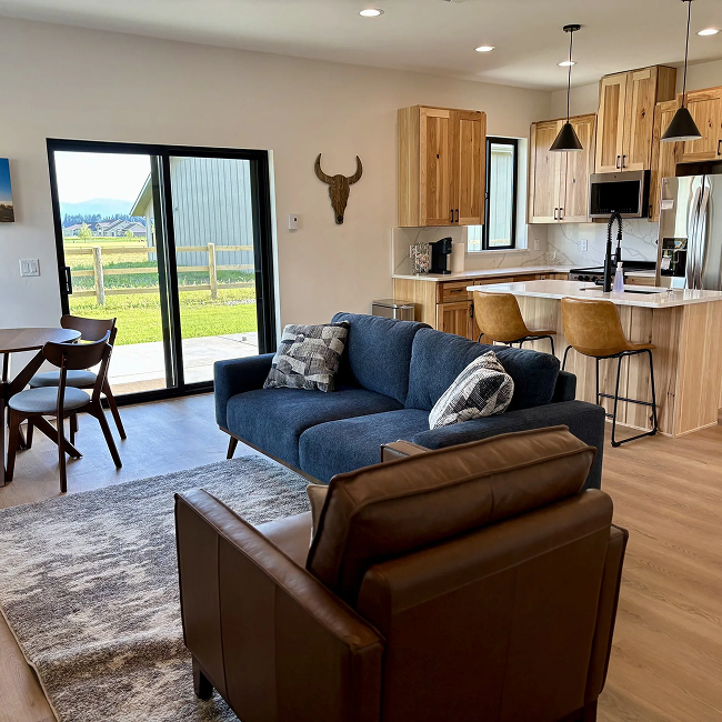 Open-concept living room and kitchen with sliding glass door leading outside, wooden cabinets, a blue sofa with pillows, a brown leather chair, and a view of green fields and mountains outside.