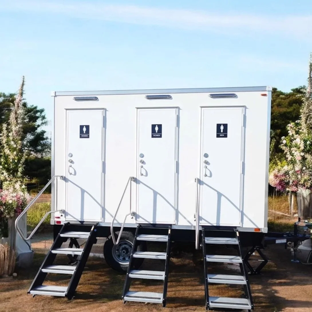 Portable restroom trailer with separate sections for women's and men's restrooms, each accessible by small stairs, set outdoors on grassy ground with trees and bushes nearby.