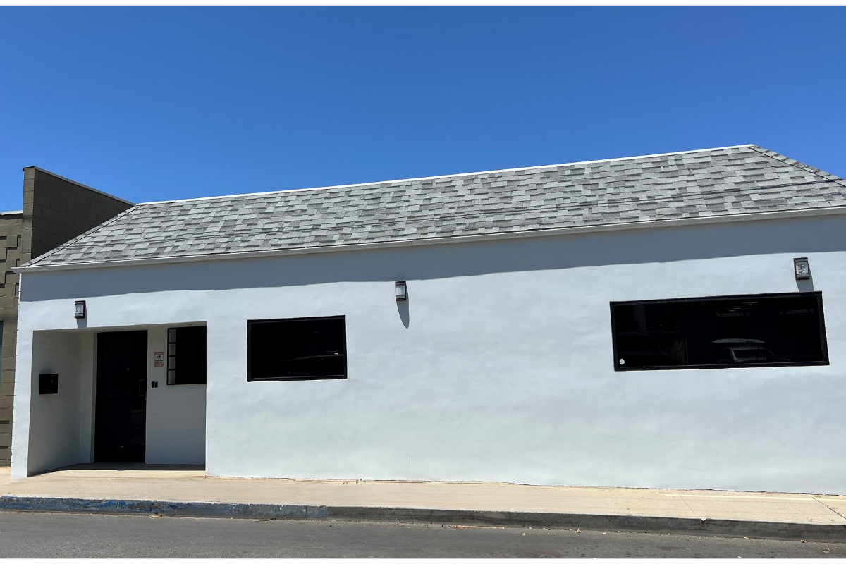 A white biorepository building with two horizontal black windows and a black door, under a gray shingle roof, against a bright blue sky.