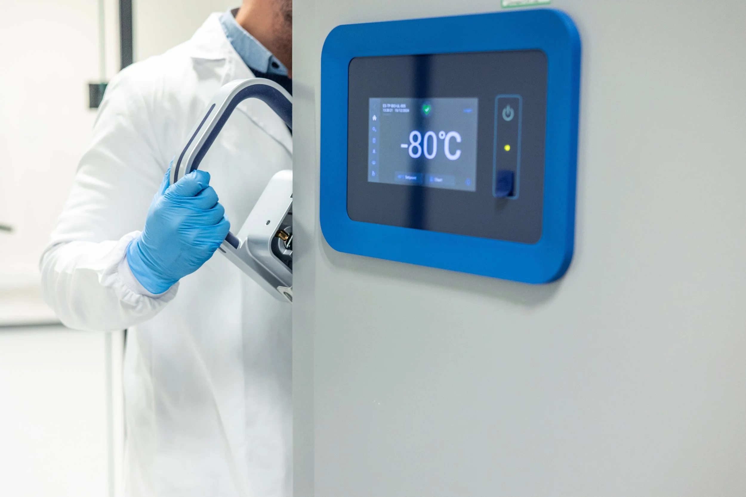 A scientist in a white lab coat and blue gloves opens a laboratory freezer, displaying a temperature of -80°C.
