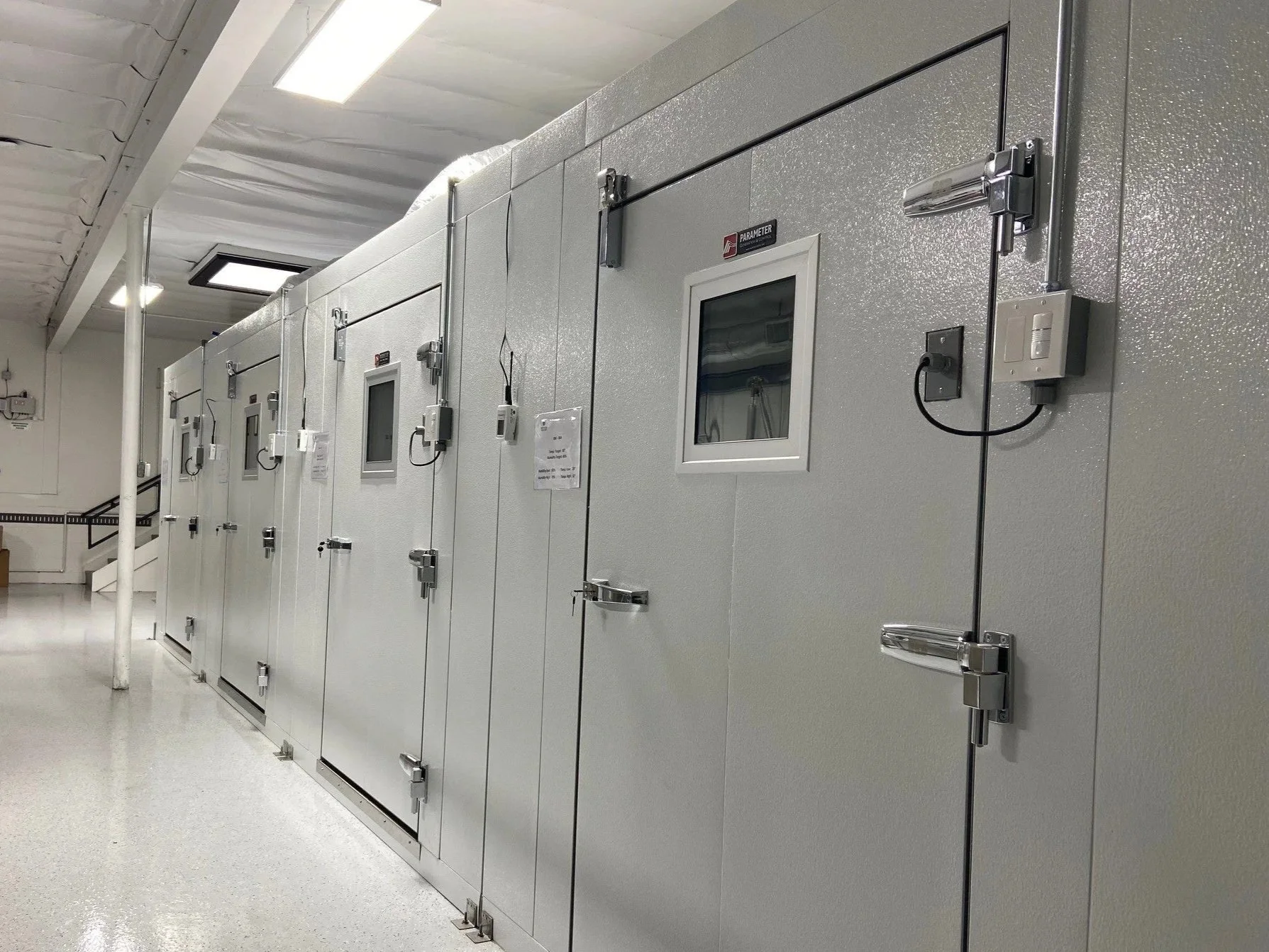 A row of large, white walk-in stability chambers with small viewing windows and secured doors in a clean, well-lit biorepository