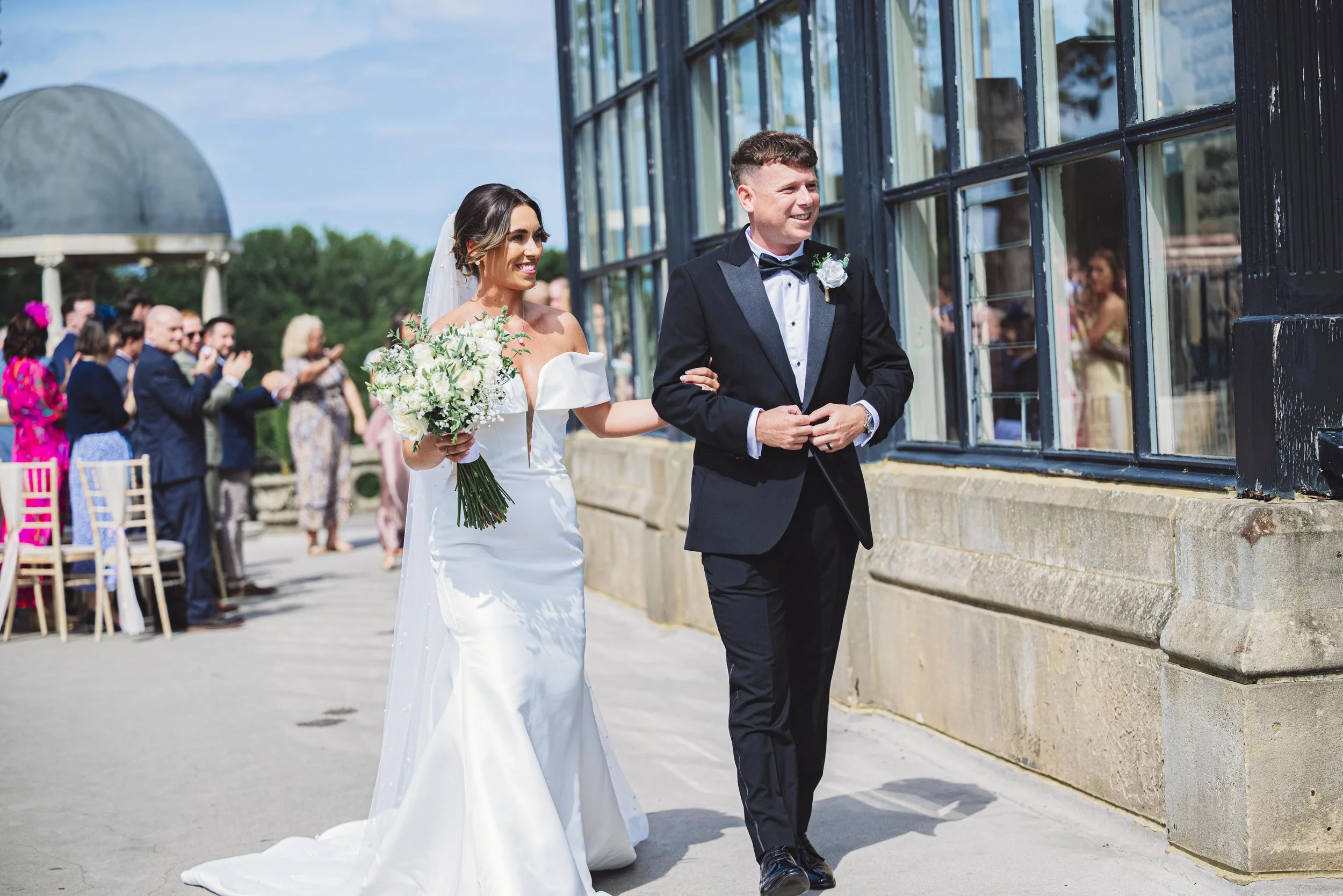 Bride and groom walking outdoors at a wedding ceremony, with the bride holding a bouquet of white flowers and the groom in a black tuxedo, guests clapping in the background.