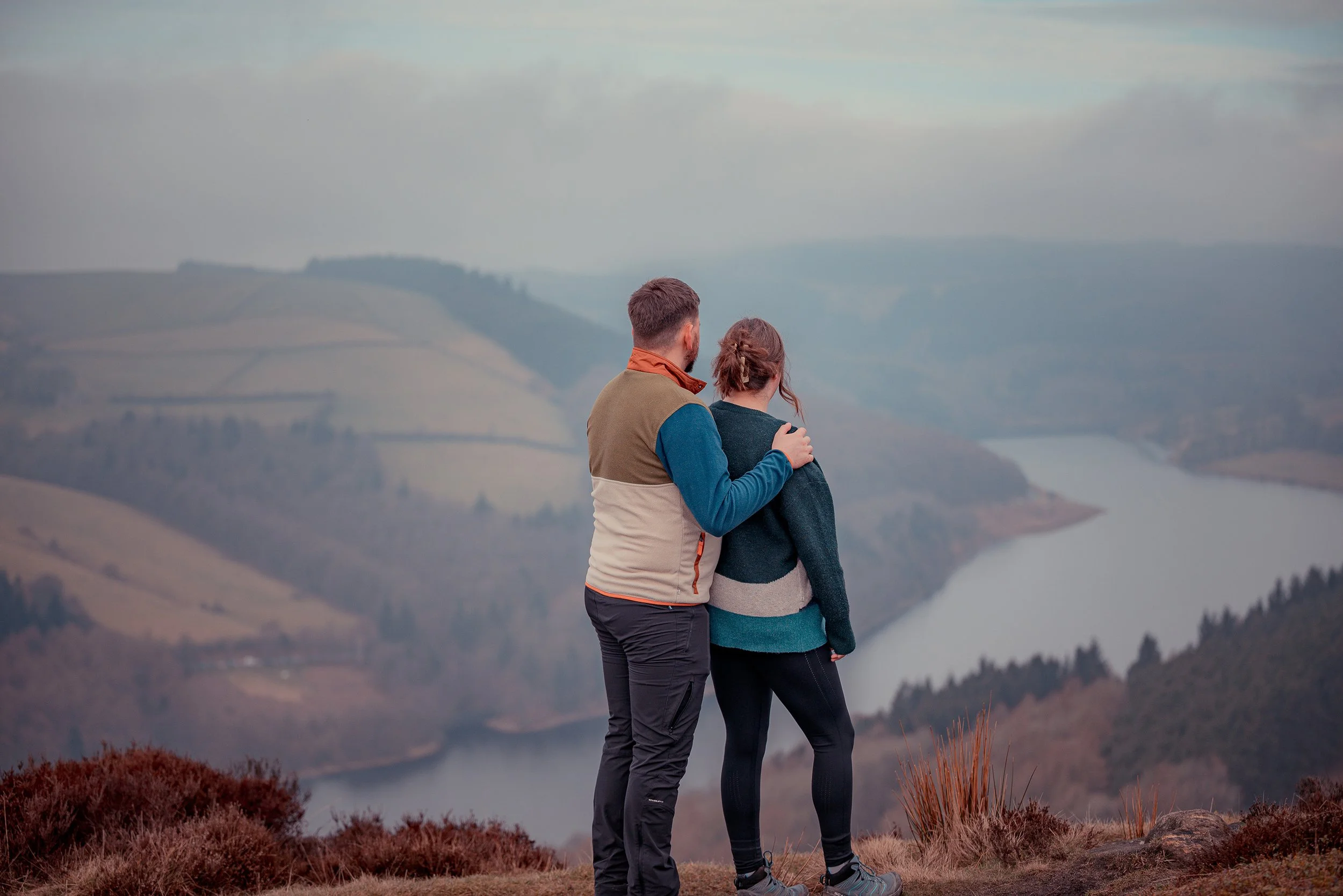 A man and woman stand together on a mountain overlooking a lake and rolling hills, with the man placing his hand on the woman's shoulder.