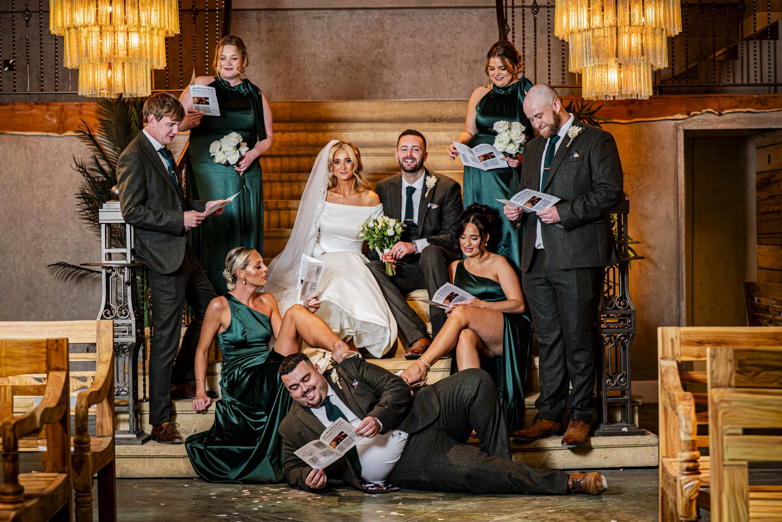 A wedding party with the bride and groom sitting on stairs surrounded by bridesmaids and groomsmen, all wearing formal attire, holding programs, in an indoor venue with wooden accents and warm lighting.