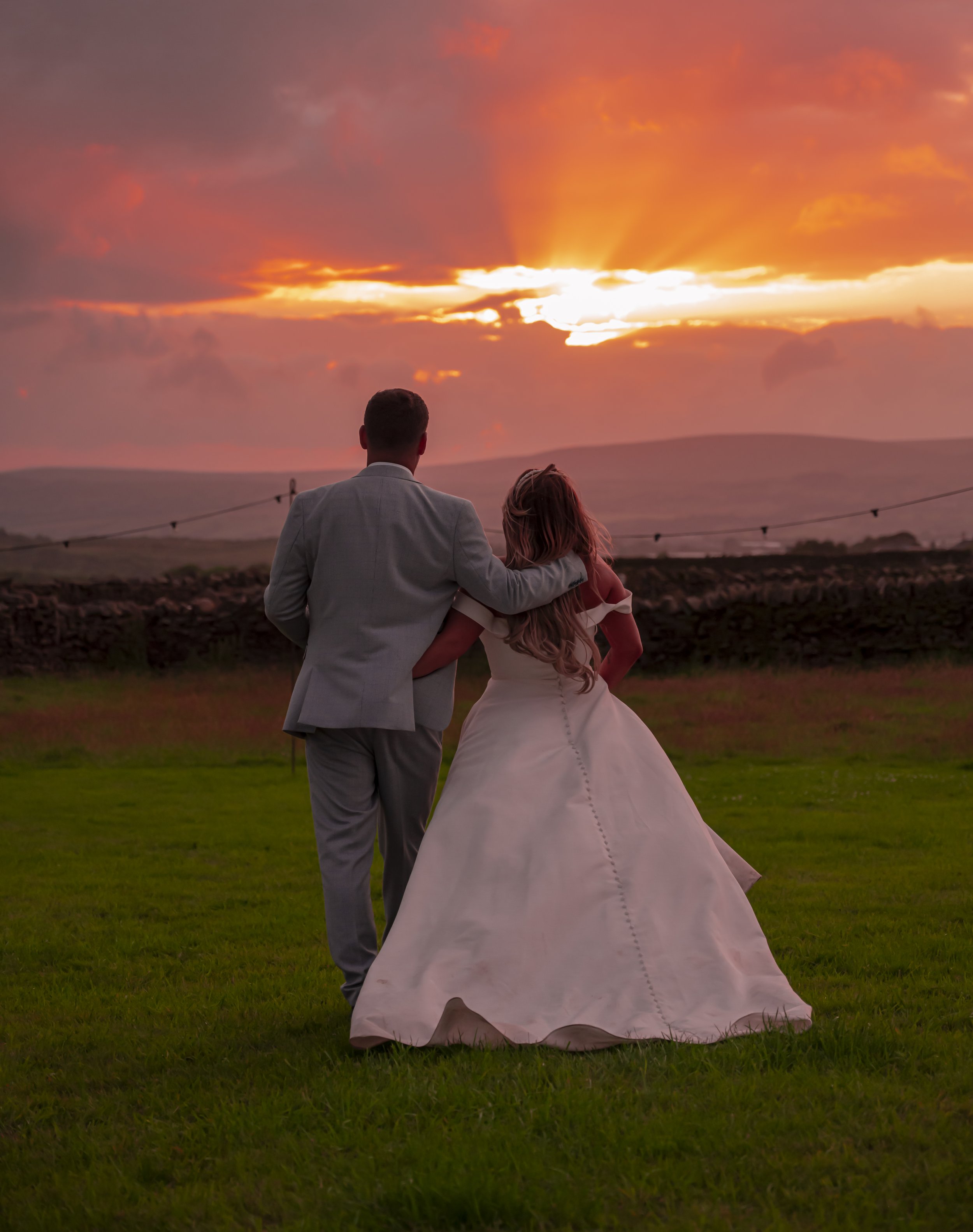 A couple dressed in wedding attire walking on grass during sunset, with the man in a gray suit and the woman in a white wedding gown, holding each other, and a dramatic sky with orange and pink hues in the background.