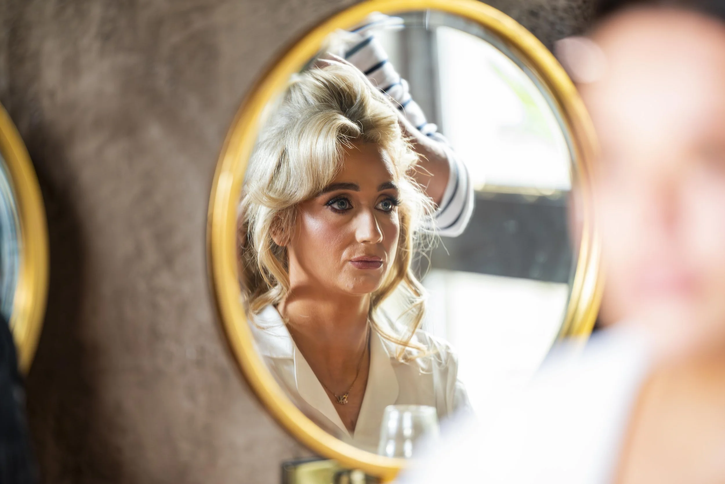 Woman with blonde hair looking into a mirror, with a stylist behind her adjusting her hair.