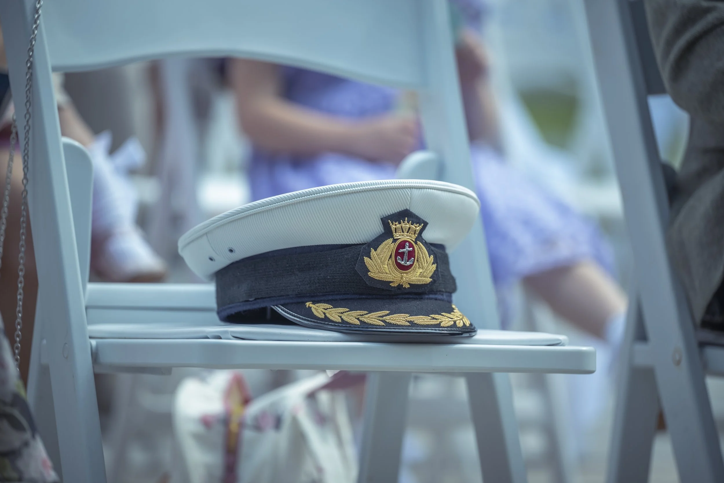 A white and navy blue naval officer's cap on a white chair, with children in formal dresses in the background.