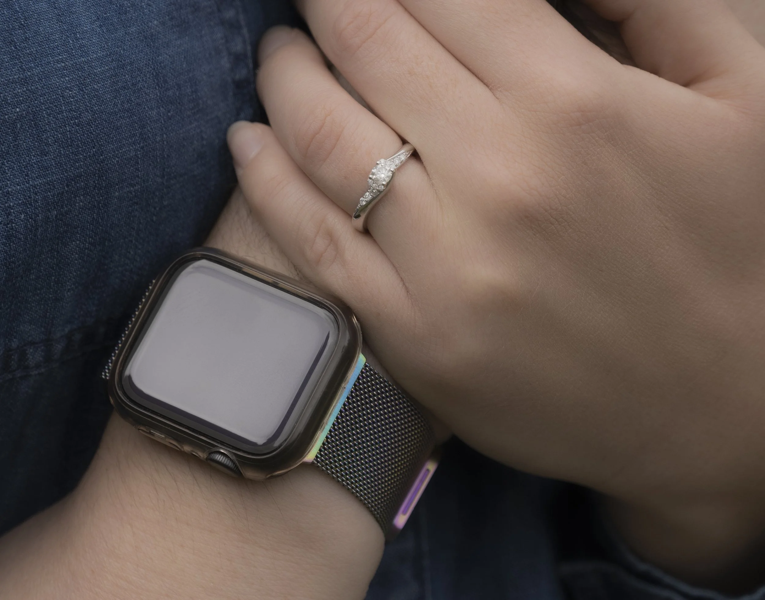 A close-up of a person's left hand wearing a silver ring with small diamonds, a smartwatch with a black band, resting on another person's arm.