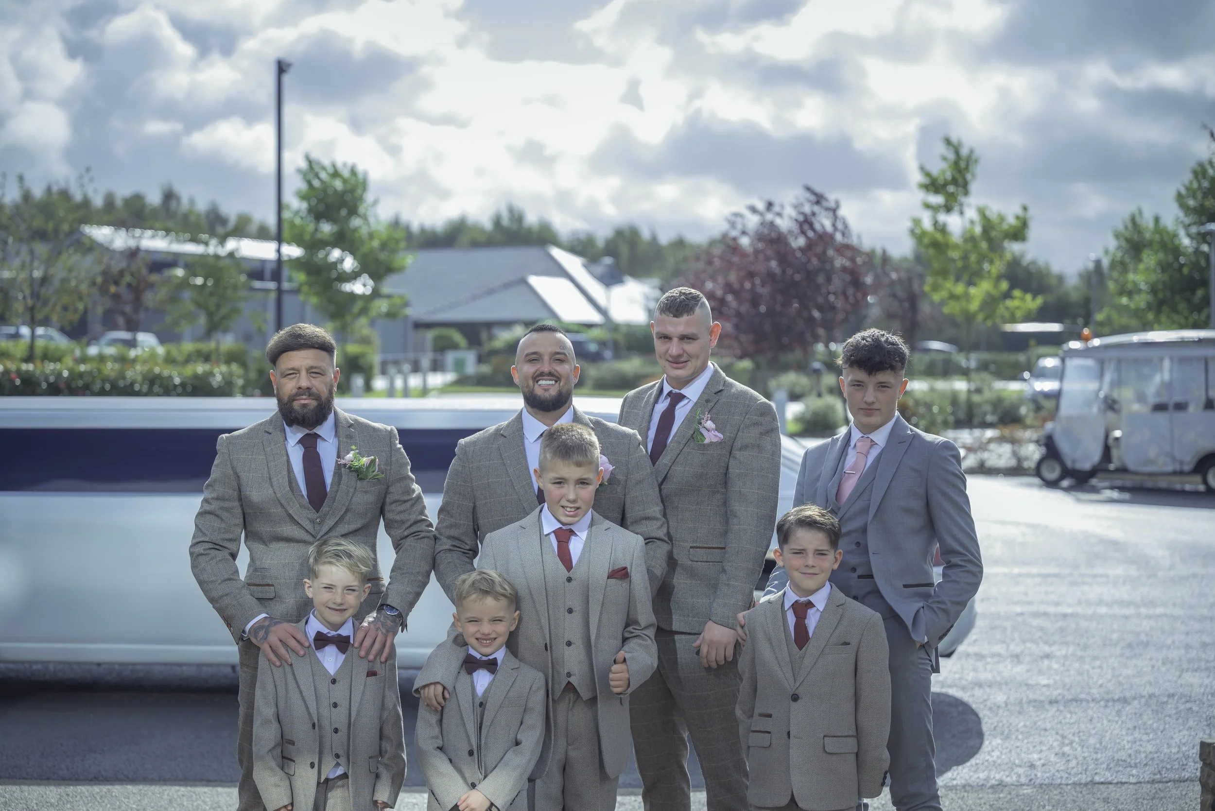 A group of men and boys in suits, standing outdoors in a parking lot on a cloudy day, smiling for a photo.