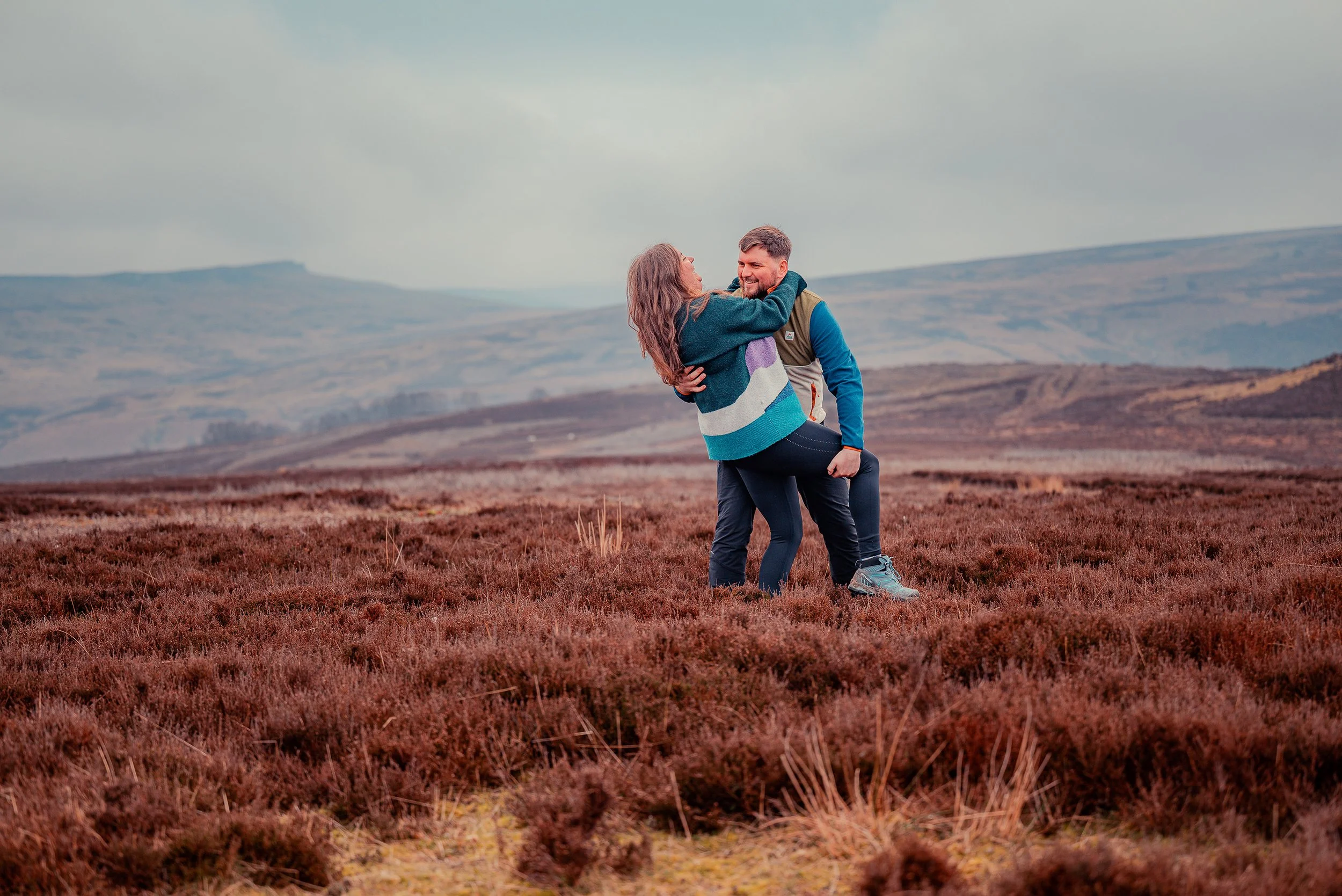 A couple smiling and embracing in a field of brown shrubs with rolling hills in the background under a cloudy sky.
