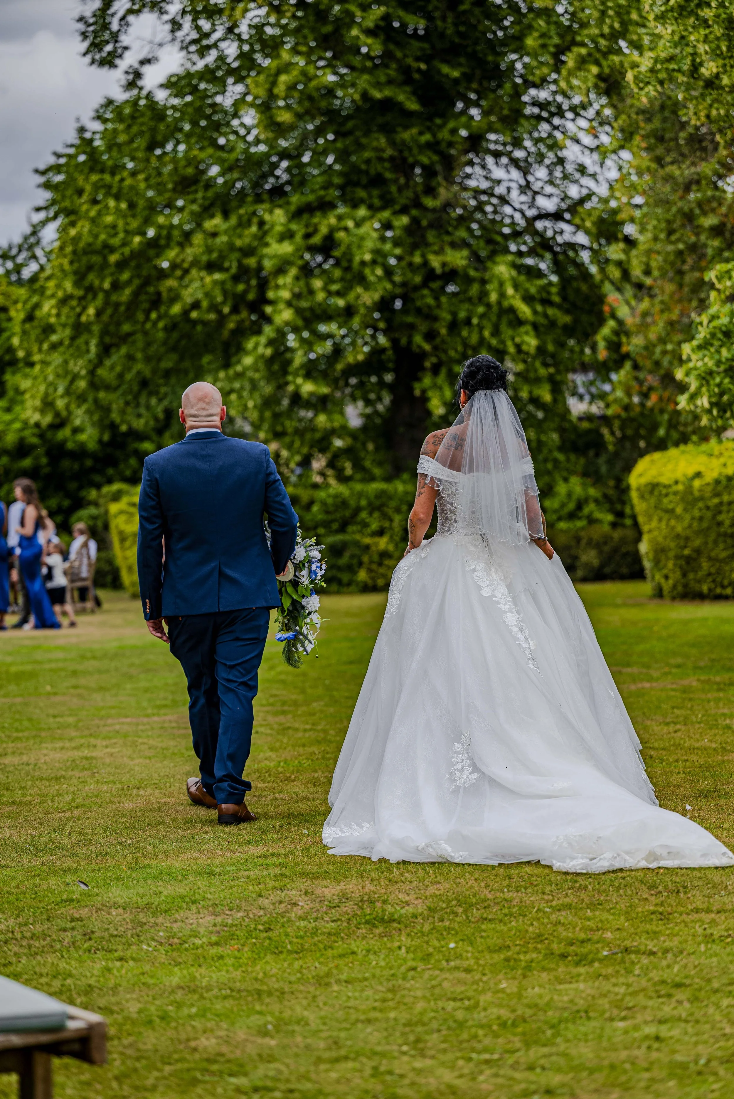 A bride in a white wedding gown and veil walking away with a groom in a blue suit in a garden setting during a wedding ceremony.