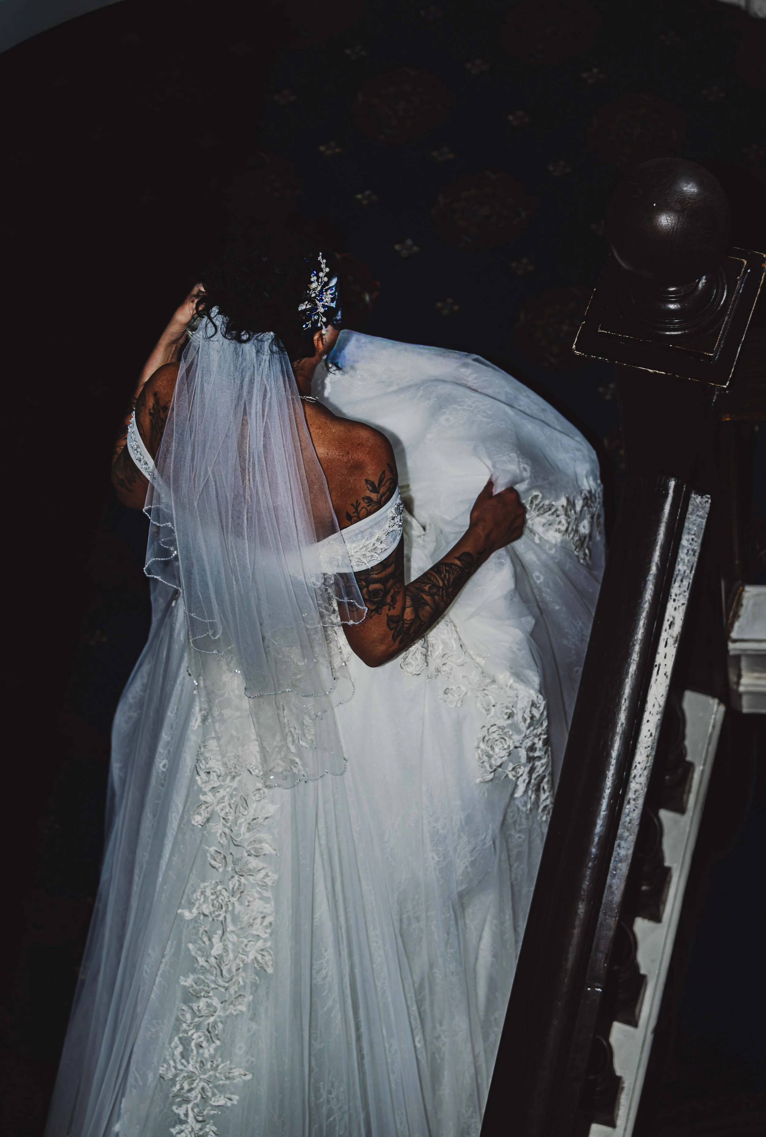 A bride in a white wedding dress and veil adjusting her gown on a staircase, showing her tattoos on her arm and shoulder.