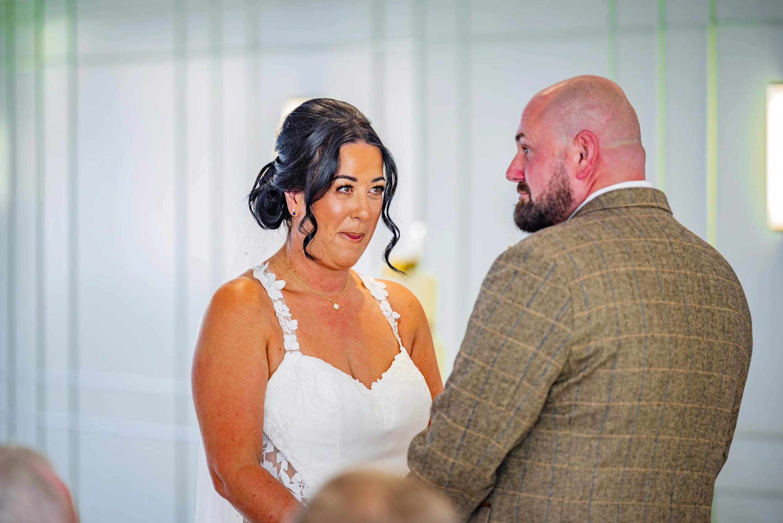 A woman and a man are having a wedding ceremony indoors. The woman is wearing a white dress with floral straps, and the man is dressed in a brown plaid suit. They are standing close together, looking at each other.