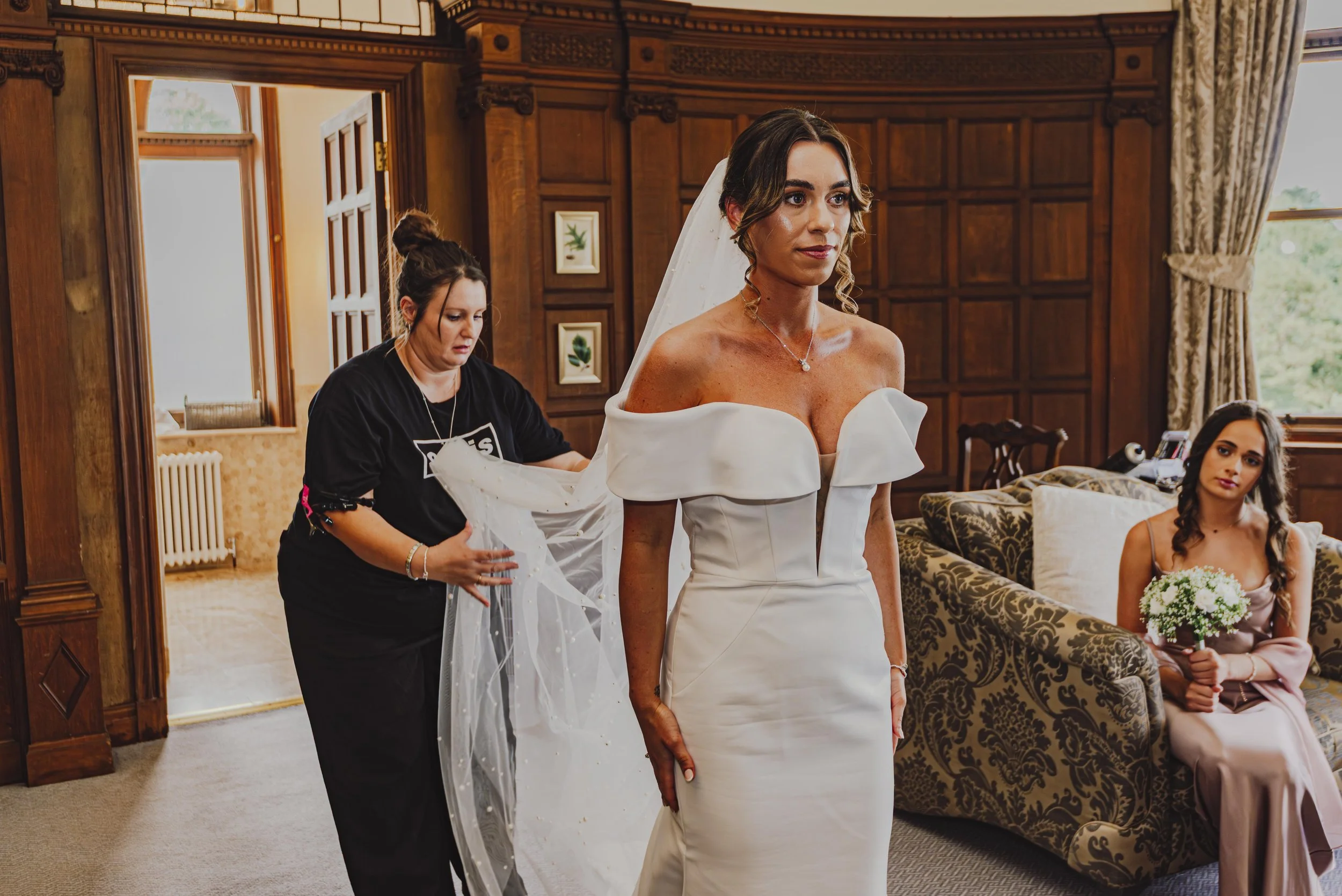 A bride in a white off-shoulder wedding gown being assisted with her veil by a woman in black. Another woman in a beige dress is sitting on a patterned sofa holding a bouquet of white flowers.