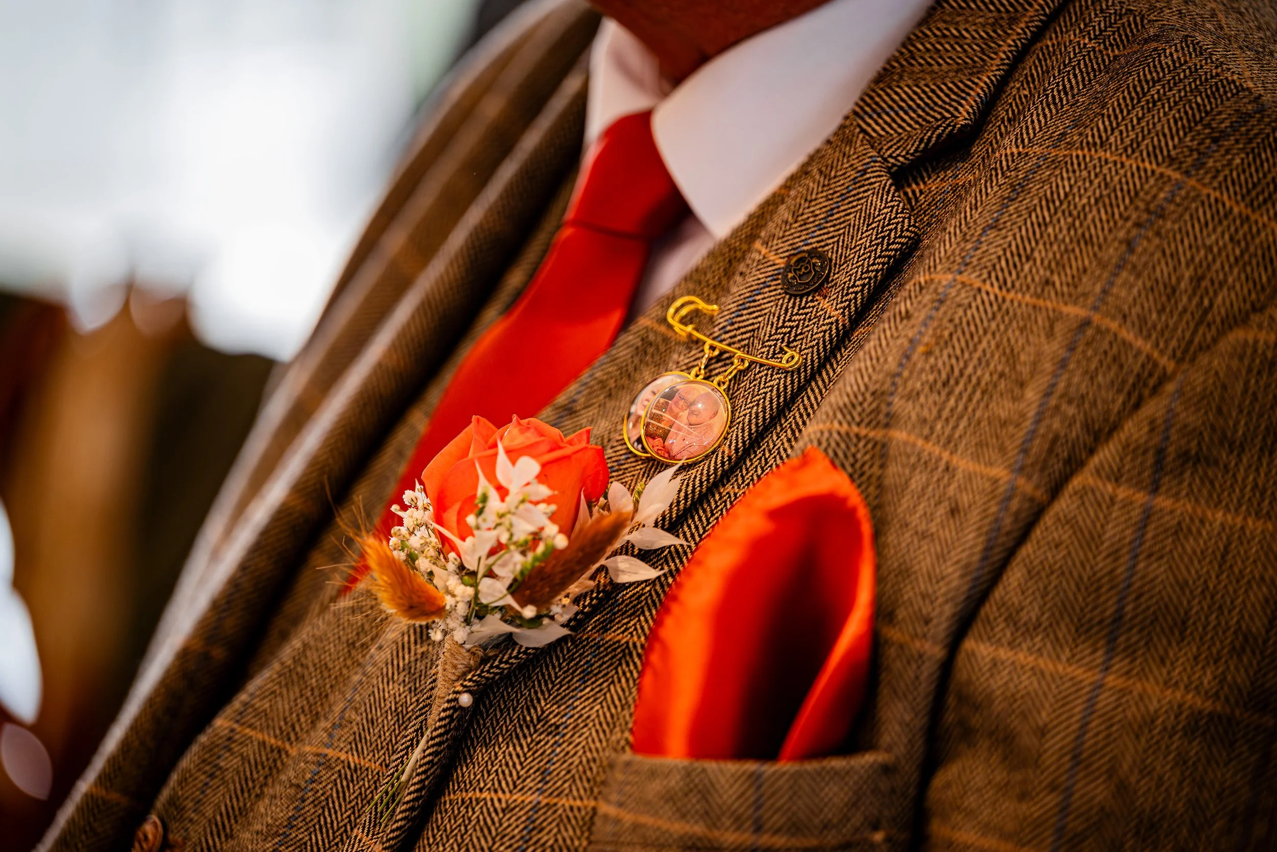 Close-up of a man in a brown plaid suit with an orange pocket square, red tie, and a boutonniere featuring a peach rose with small white flowers and foliage, along with a gold lapel pin.