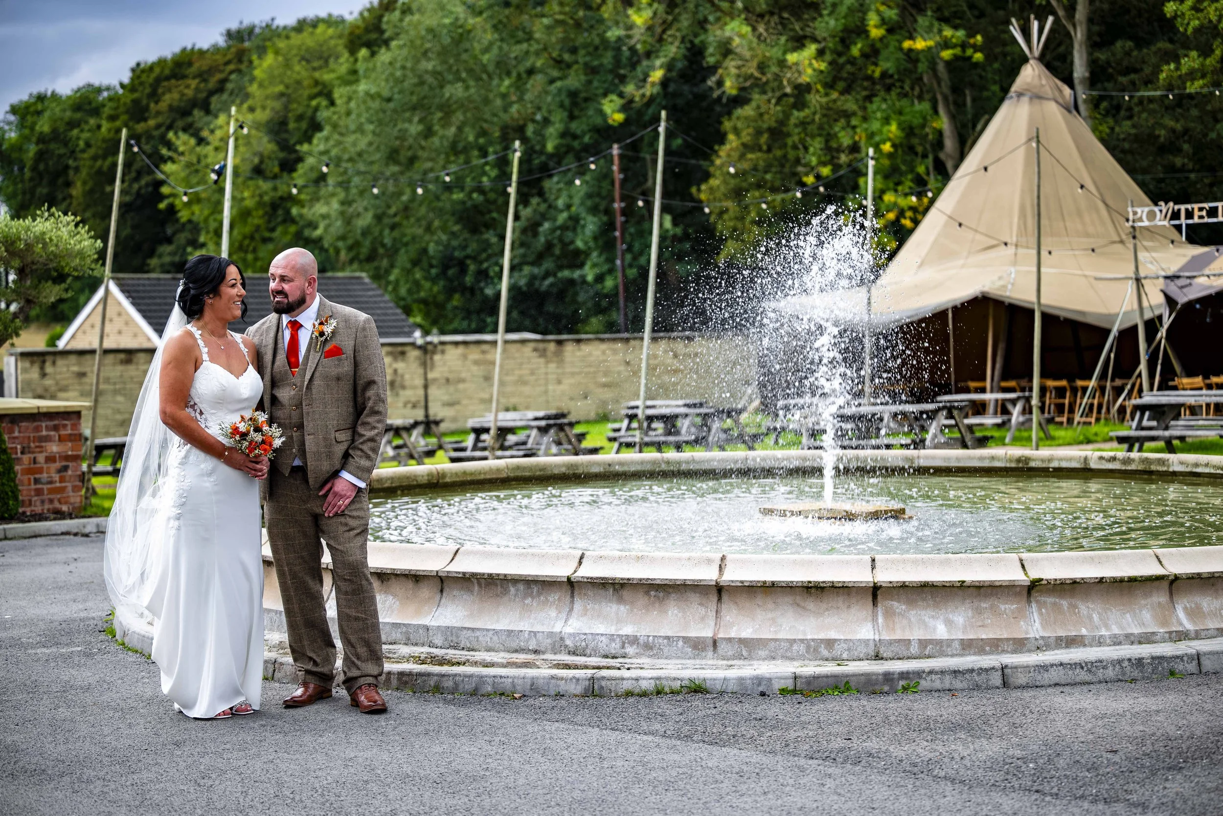 A bride and groom in wedding attire standing near a fountain outdoors, smiling and looking at each other.