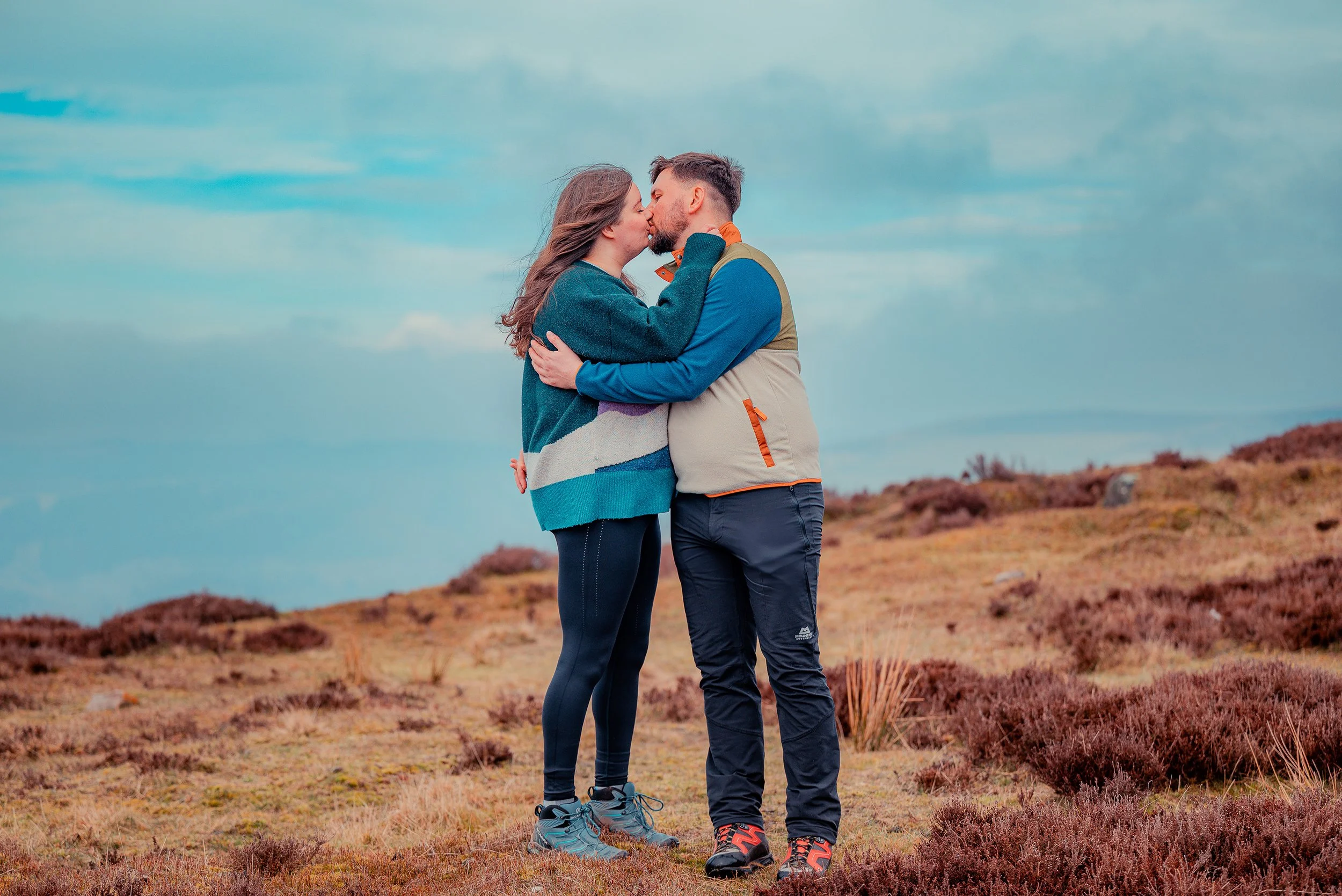 A couple kissing outdoors on a hilly landscape with sparse bushes, dressed in outdoor gear.