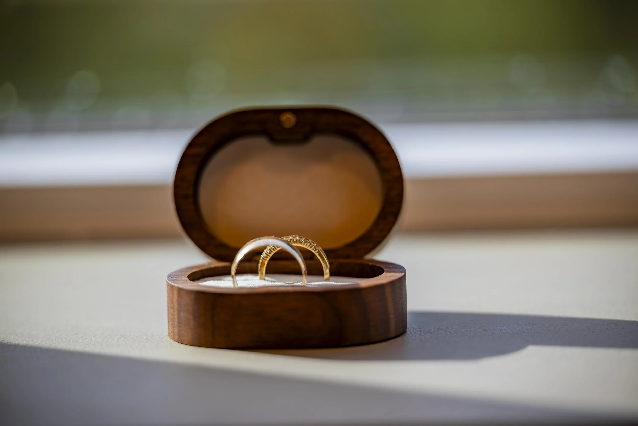 Gold wedding rings in a wooden ring box, placed on a white surface near a window with blurred outdoor scenery.