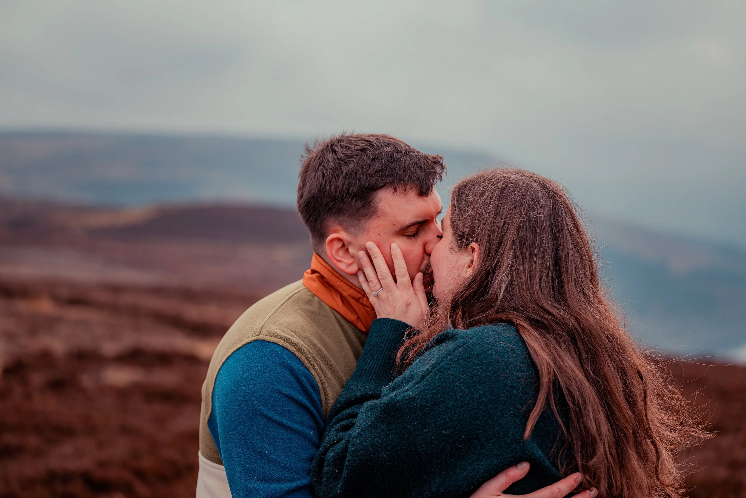 A couple is sharing a kiss outdoors on a cloudy day with mountains in the background.
