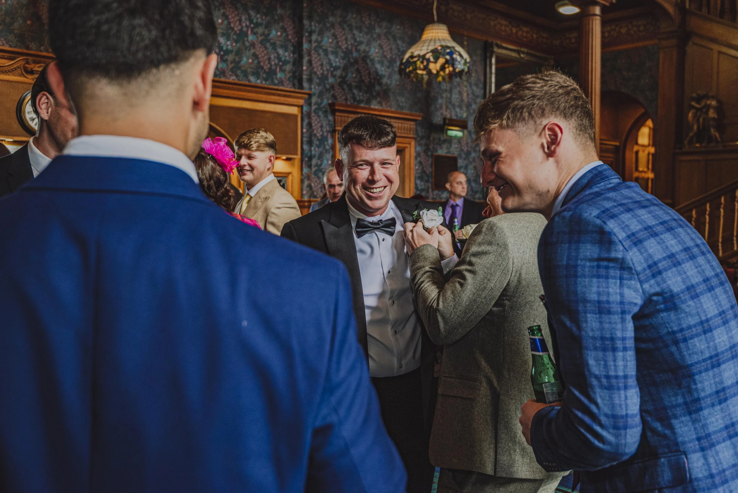 A group of people at a wedding reception, with the focus on a smiling groom in a tuxedo, as he interacts with others holding drinks and engaging socially in a decorated wood-paneled room.